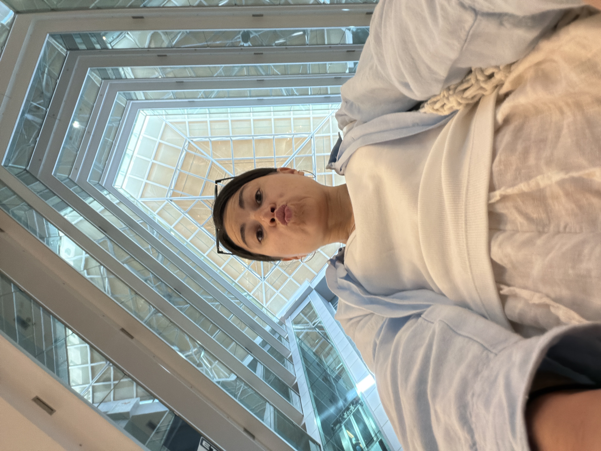 A low-angle shot shows a woman taking a selfie in what appears to be an atrium. The background shows a multi-story building with glass windows and a geometrical shaped glass roof. The woman is wearing light-colored clothing, and her face is puckered in a kissing expression. The location appears to be a building with a modern design. No specific information suggests this is Budva, Montenegro.