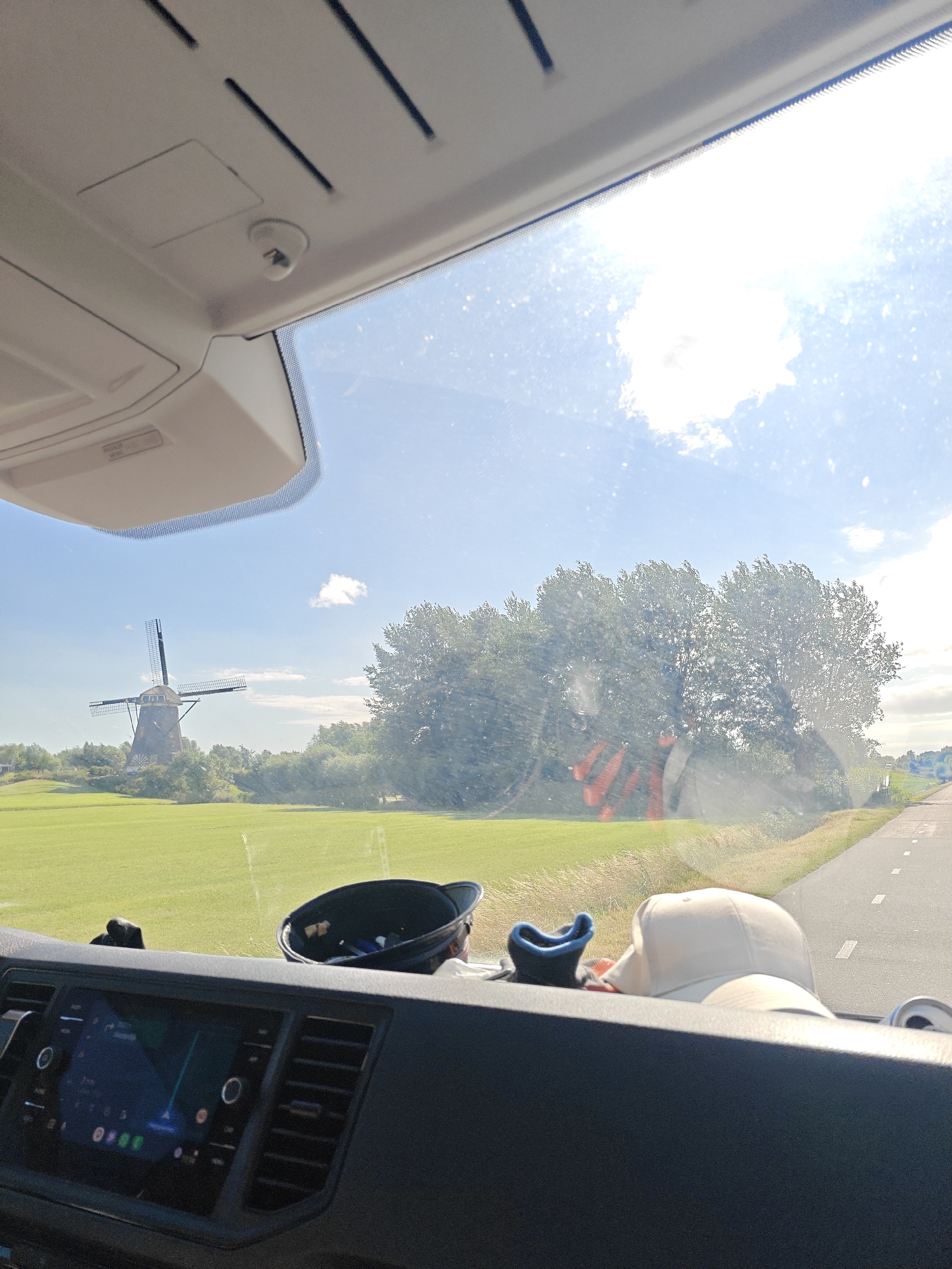 The image captures a view from inside a vehicle, looking out through the windshield on a sunny day. The scene depicts a rural landscape in Leimuiden, Netherlands, with a traditional Dutch windmill visible in the distance. The windmill has dark sails and is situated on a small mound surrounded by lush green fields. A line of trees with full foliage borders the field and extends towards the right side of the image.
In the foreground, the dashboard of the vehicle is visible, with a touchscreen display showing a navigation interface. Various objects are placed on the dashboard, including a black cap with a blue lining, a white baseball cap, and what appears to be a small can. The sky is blue with scattered white clouds, and the sunlight suggests it is daytime, likely mid-morning to late afternoon. The road is visible to the right, with white markings indicating lanes. The overall impression is one of a pleasant, scenic drive through the Dutch countryside. There is no visible text in the image.