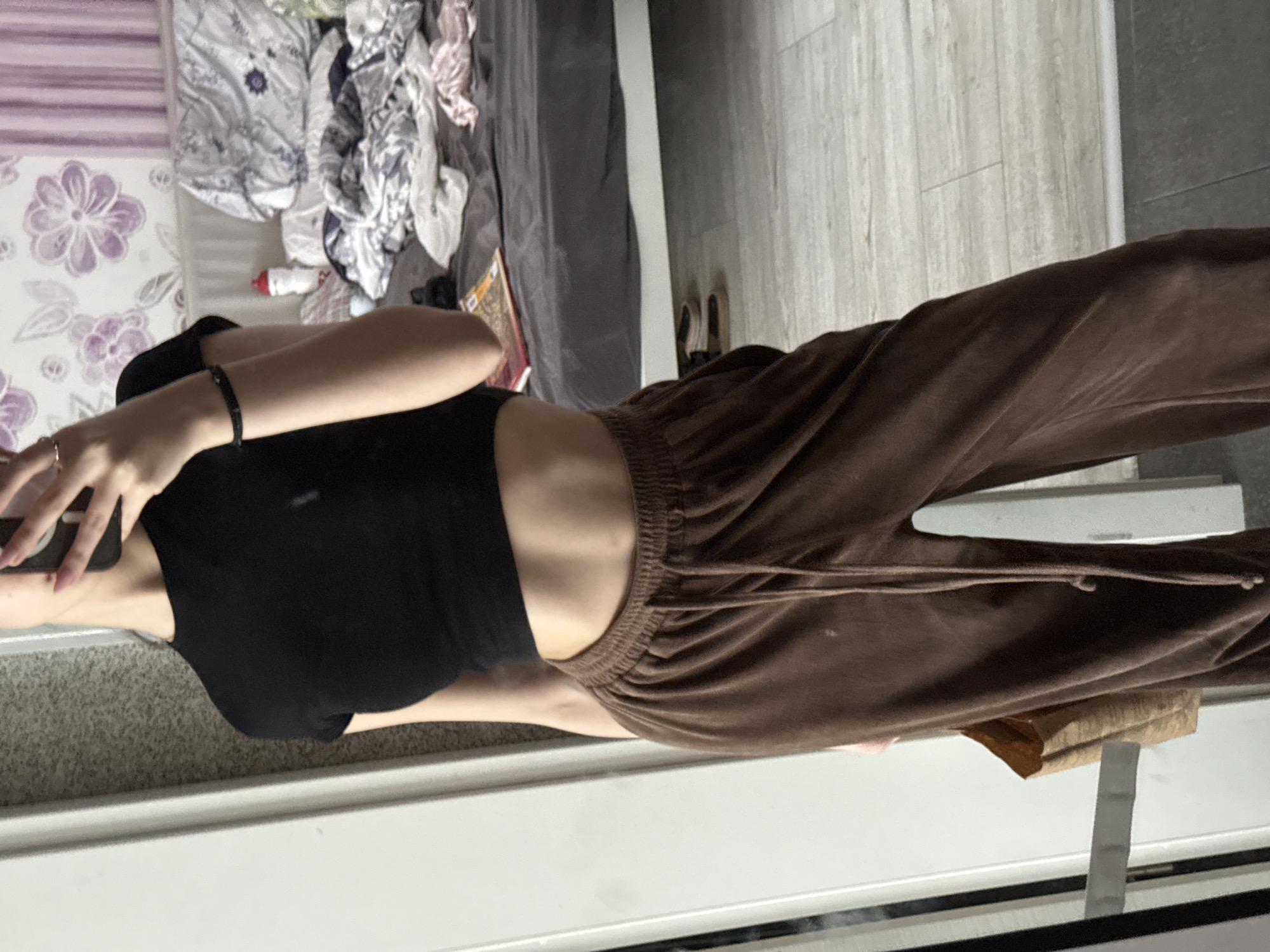 A person is standing indoors, likely in an apartment. The person is wearing a black crop top and brown sweatpants. The background shows a messy bed, part of the floor, and a white wall. The location appears to be indoors. It is not possible to identify if the location is in Sofia, Bulgaria based on this image.