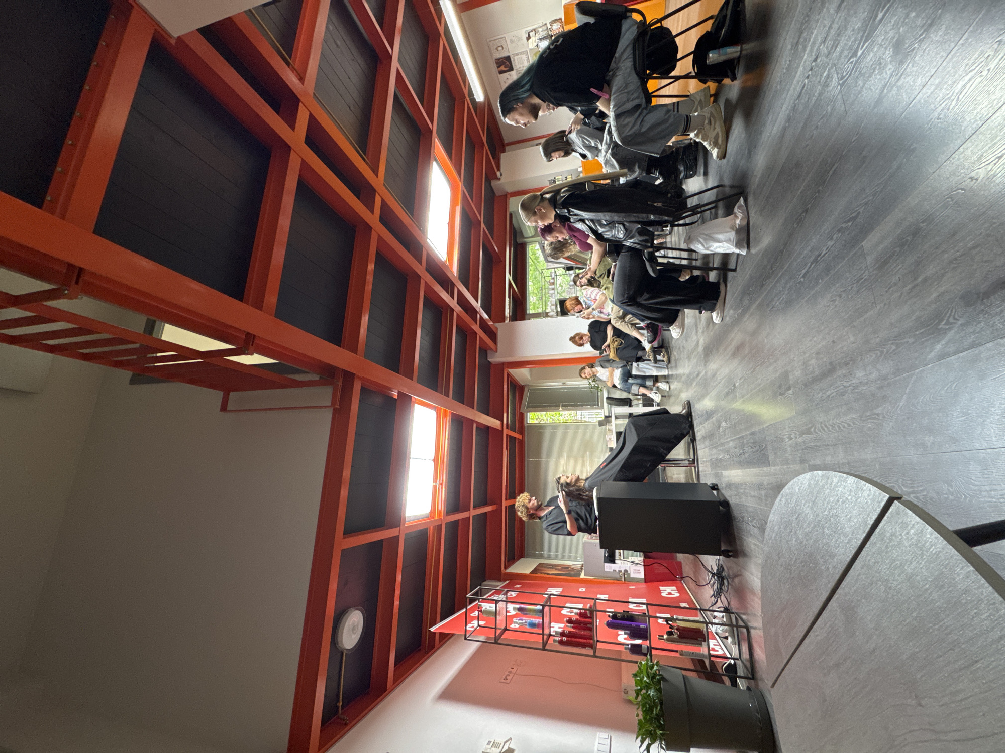 The image depicts the interior of a hair salon in Tbilisi, Georgia, characterized by a modern architectural design with exposed orange metal framework. Along one side, clients are seated in a row, receiving haircuts or styling. The scene is populated with people in various stages of hairstyling; some clients are draped with protective capes. Salon equipment, including hair dryers, styling tools, and a product display, is visible. The salon has grey wooden flooring. A round table is situated near a workstation where a client is receiving service. A plant is in a pot near the tool display. The overall impression is that of a bustling and trendy hair salon in the city.