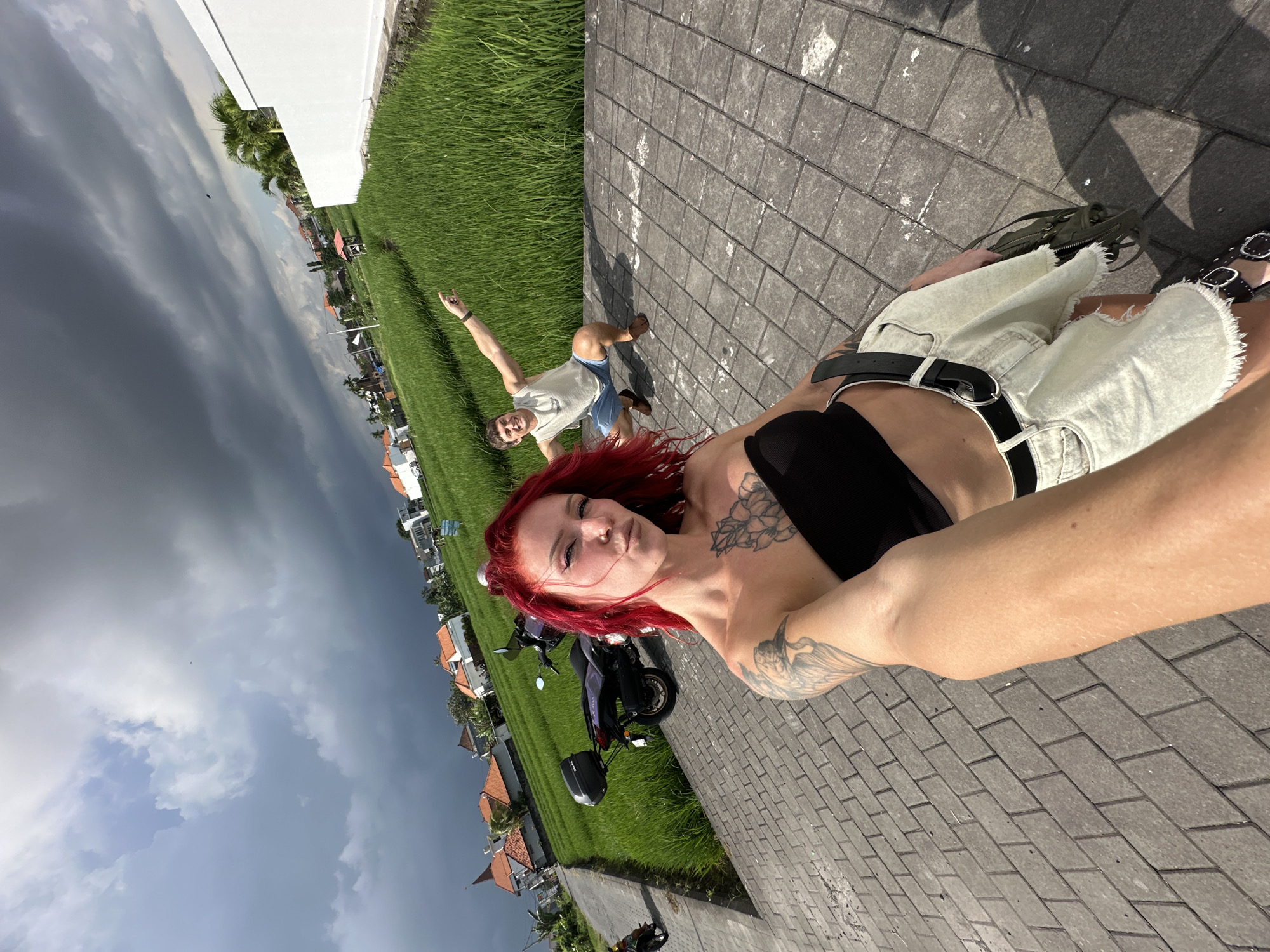 The image is a rotated selfie taken on a paved surface in North Kuta, Indonesia. A person with red hair, tattoos on their shoulder and arm, a black top, light-colored shorts, and a black belt is in the foreground. Their arm extends to hold the camera. In the background, another person is lying in a rice field, with their arm raised. Buildings with orange roofs are visible in the distance. A motorcycle is parked on the side of the road. The sky is cloudy and dark.