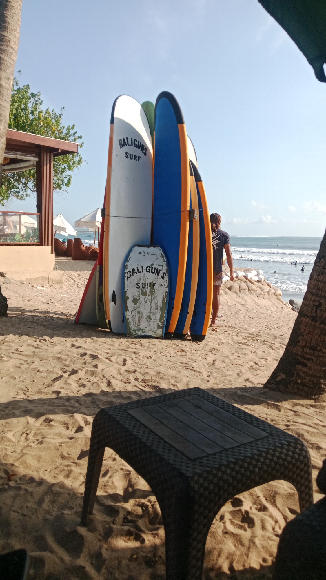 This image depicts a beach scene in Kuta, Indonesia. In the foreground, a dark, woven plastic table sits on the sand, casting a shadow. Behind the table, a collection of surfboards are propped upright against each other. The surfboards are of various colors, including white with "BALIGUNS SURF" written on one, blue with orange stripes on others, and a green surfboard. A small, weathered board with "BALI GUN.S SURF" written on it leans in front of the larger surfboards. In the middle ground, a person wearing a dark t-shirt and shorts stands facing the ocean, their back to the camera. Beyond them, the ocean stretches to the horizon with gentle waves. Sandbags are visible on the beach near the water. In the background, to the left, part of a wooden structure with a roof, possibly a beachside cafe or shelter, is visible, with some people sitting under it. Palm trees frame the scene on the left and right. The sky is clear and blue.