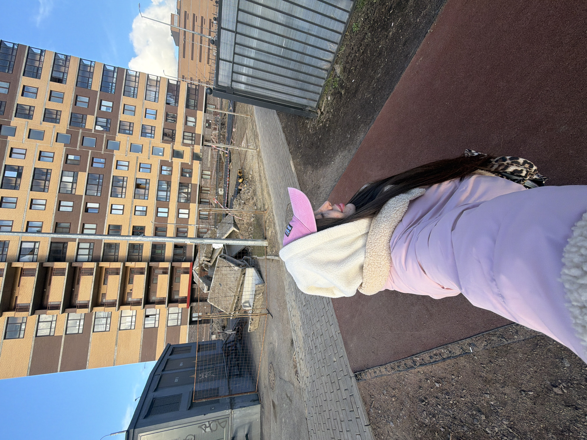 A woman wearing a pink baseball cap, a white hooded sweatshirt, and a light purple jacket lies on a brown and grey tiled area, possibly a sidewalk or path. She faces towards the building on the left.
In the background, there's a tall residential building with a tan and brown facade, featuring rows of windows. There are signs of construction or demolition in the middle ground. A grey metal structure, possibly a fence or shed, stands between the woman and the construction site. The sky is blue with scattered clouds.
The buildings and presence of construction suggest an urban location, possibly Saint Petersburg, Russia, based on building architectural styles and construction practices.