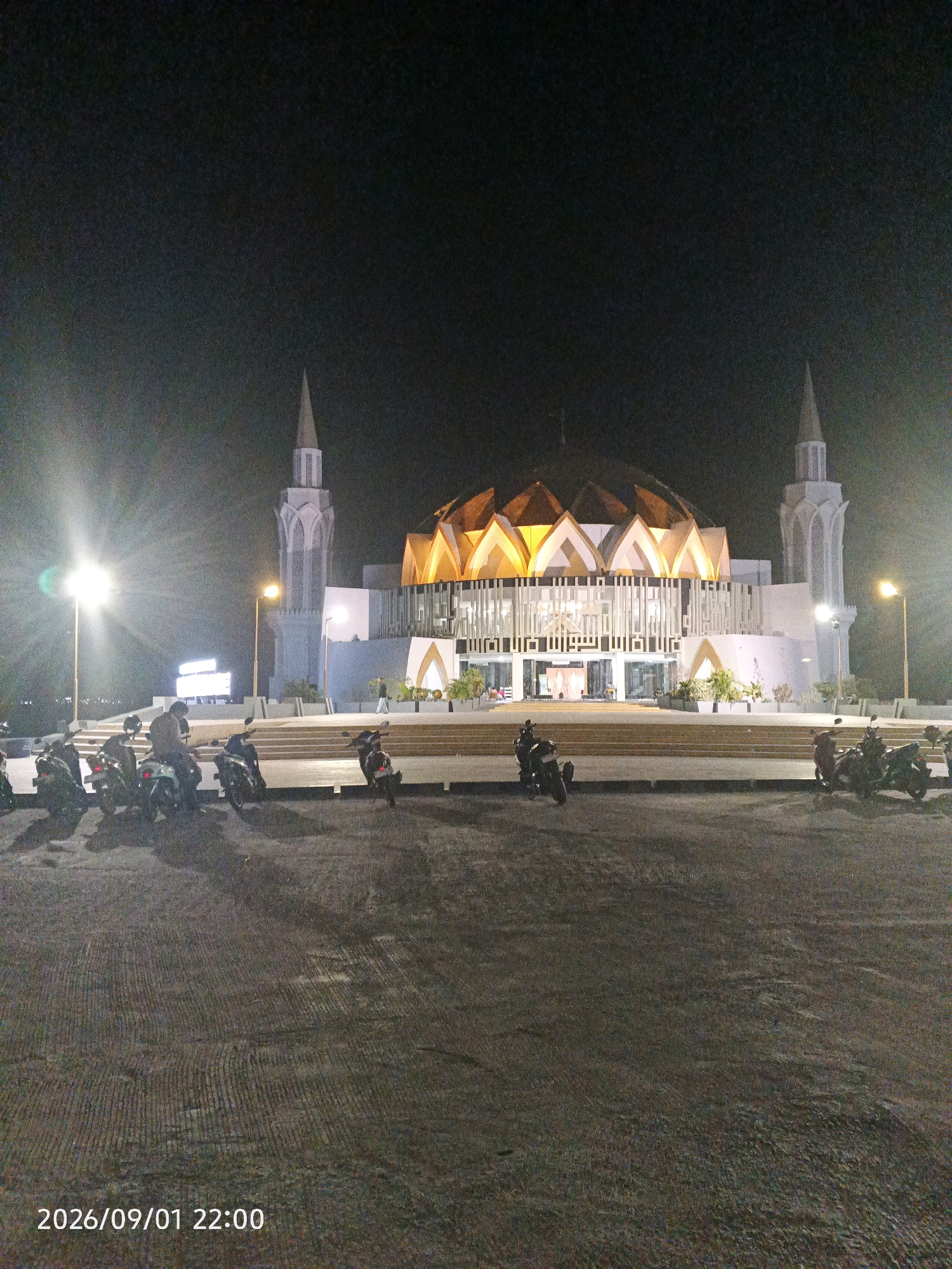 The image shows a night view of the Grand Mosque of Al-Manar in Pare-Pare, South Sulawesi, Indonesia. The mosque features a distinctive modern architectural design with a domed roof structure illuminated from within by warm, golden light, and two tall, slender minarets on either side. The exterior of the mosque is primarily white, with a patterned facade. In the foreground, a paved area is visible with several motorcycles parked. A street light with a bright glow is to the left of the frame, and another street light is positioned to the right. The sky is dark, indicating it is nighttime. In the bottom left corner, a timestamp reads "2026/09/01 22:00". There are a few indistinct figures of people on the steps leading up to the mosque and near the parked motorcycles. The overall scene is one of a well-lit, prominent religious building at night.