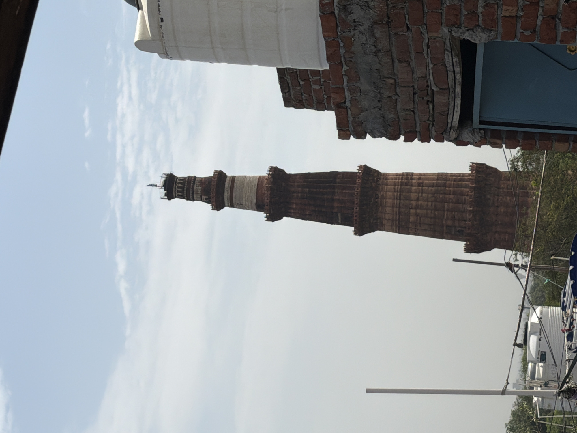 The image shows the Qutub Minar, a UNESCO World Heritage Site, in Delhi, India. The tower, made of red sandstone, is tall and slender with multiple stories. In the foreground, there is a partially visible water tank and a brick structure. The sky is clear with some scattered clouds. The overall scene appears to be a view of the Qutub Minar from a nearby rooftop.