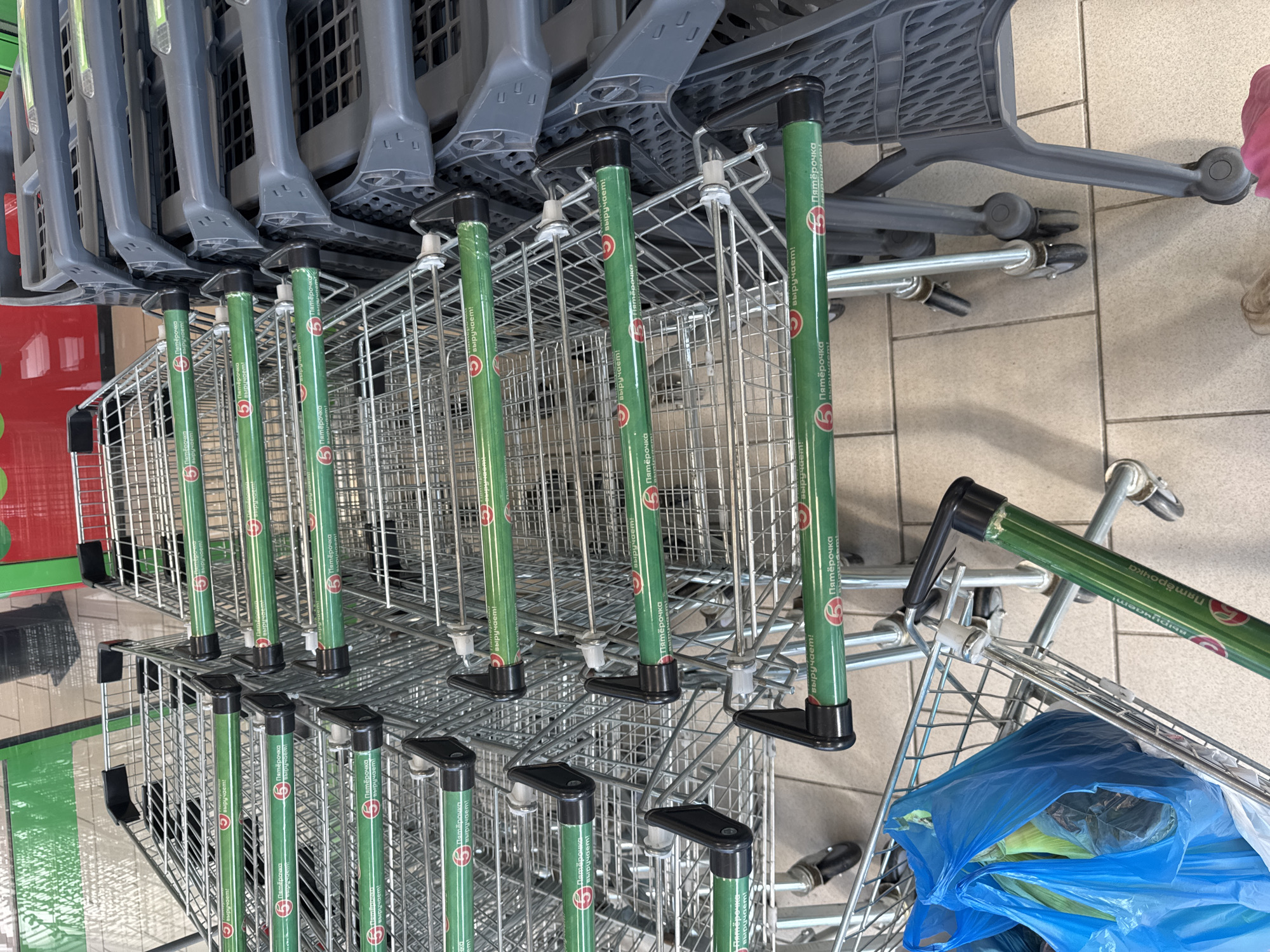 This image shows a row of stacked shopping carts in what appears to be a grocery store or a similar retail environment. The carts are made of metal wire, with green handles that have the "Pyaterochka" logo and text in Russian. Some of the carts are nested inside each other, a common way to store them when not in use. In the foreground, there is a blue plastic bag filled with items, placed inside one of the carts. The floor is tiled. The lighting suggests an indoor setting. The visible text is in Russian and appears to be branding for the "Pyaterochka" supermarket chain. The overall scene depicts a typical retail setting with organized shopping carts. There are no people visible in the image. The image is taken from a slightly elevated angle, looking down at the carts.