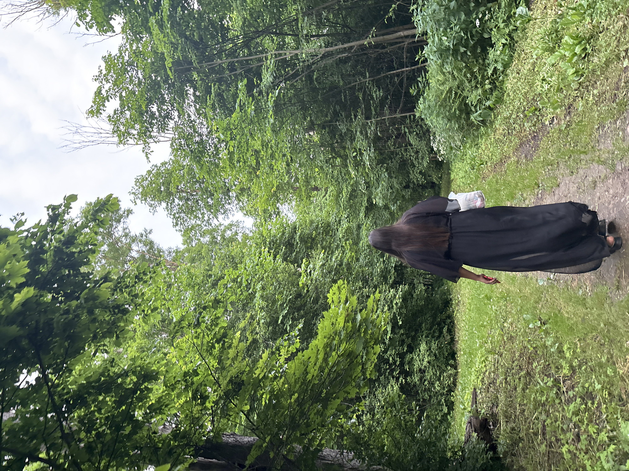 A person with long, dark hair wearing a flowing black dress walks along a dirt path through a lush, green forest. The path is bordered by grass and dense foliage. Sunlight filters through the canopy of trees, creating dappled light on the path. In their right hand, the person carries a white plastic bag. The scene appears to be in a natural environment, potentially a park or wooded area within the city of Gorki, Russia.
