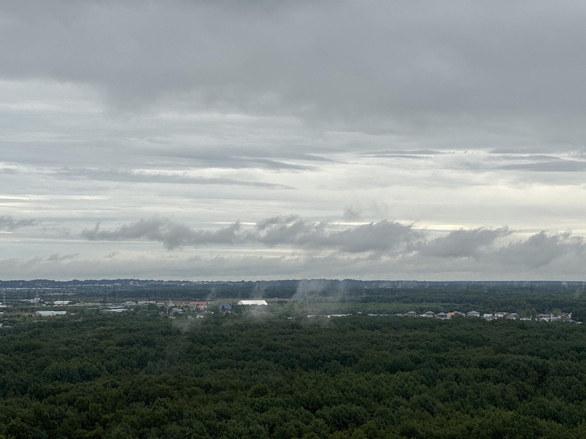 An aerial view captures a vast, dense forest of dark green trees stretching across the foreground and mid-ground. In the distance, a band of buildings, appearing to be a residential area with houses and scattered structures, is visible beyond the tree line. Further in the background, industrial or commercial buildings with white roofs and smokestacks are present, along with power lines and transmission towers. The sky is overcast with thick, grey clouds that obscure the sun, creating a diffused and muted lighting condition across the entire scene. The location is P.im.sverdlova, Russia.