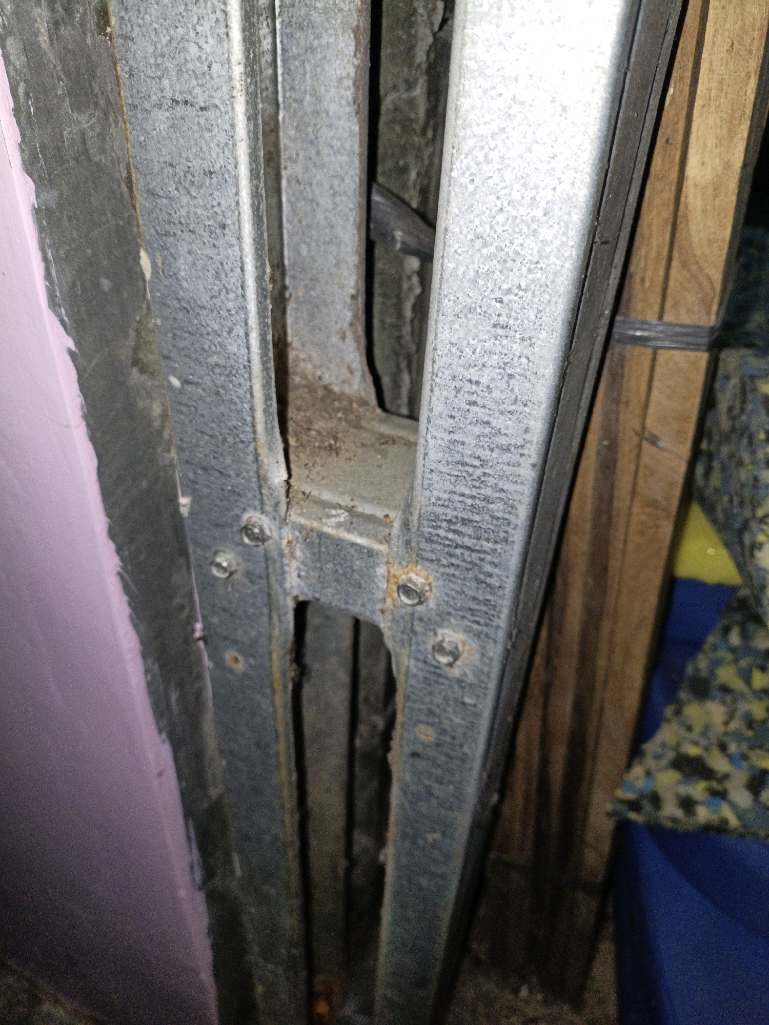The image shows a close-up view of rusty metal framing and a pink-colored surface, possibly insulation or painted wall, on the left. The metal frame appears to be part of a structure, possibly a doorway or a partition. There are visible screws attaching the metal pieces together. In the background, there are wooden planks and some colorful patterned material, possibly upholstery or padding. The scene appears to be indoors or in a sheltered area, with some debris on the ground. No people or specific Indonesian landmarks are visible. The location in the city of Cikal, Indonesia is not determinable from the image itself.