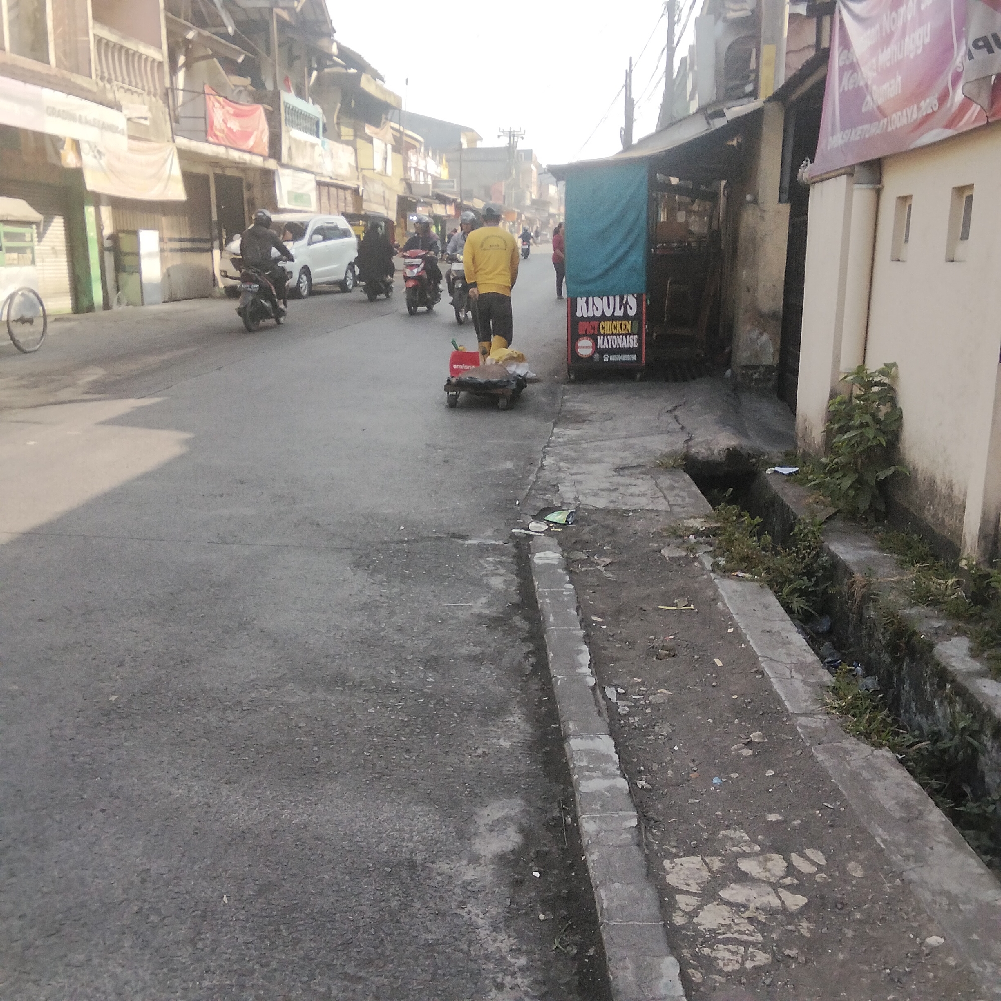 The image depicts a street scene in Cisaat, Indonesia, characterized by a weathered asphalt road with visible cracks and debris. Buildings line the right side of the street, some appearing to be shops with awnings and signage, including a prominent sign for "KISUL'S SPICY CHICKEN MAYONNAISE". Several people are present. In the foreground, a person in a yellow long-sleeved shirt and yellow boots is pushing a small, four-wheeled cart loaded with items. Behind this individual, other individuals on motorcycles are also visible, some traveling and others parked. A white car is parked further down the street, and another motorcycle is moving towards the foreground. The left side of the street shows more buildings, some with multiple stories and balconies, and banners are hanging from some of them. The overall atmosphere suggests a bustling commercial or residential area with typical street activity. A drainage ditch runs along the edge of the sidewalk on the right, with some vegetation growing in and around it. The lighting suggests it could be late morning or early afternoon, with a hazy sky.