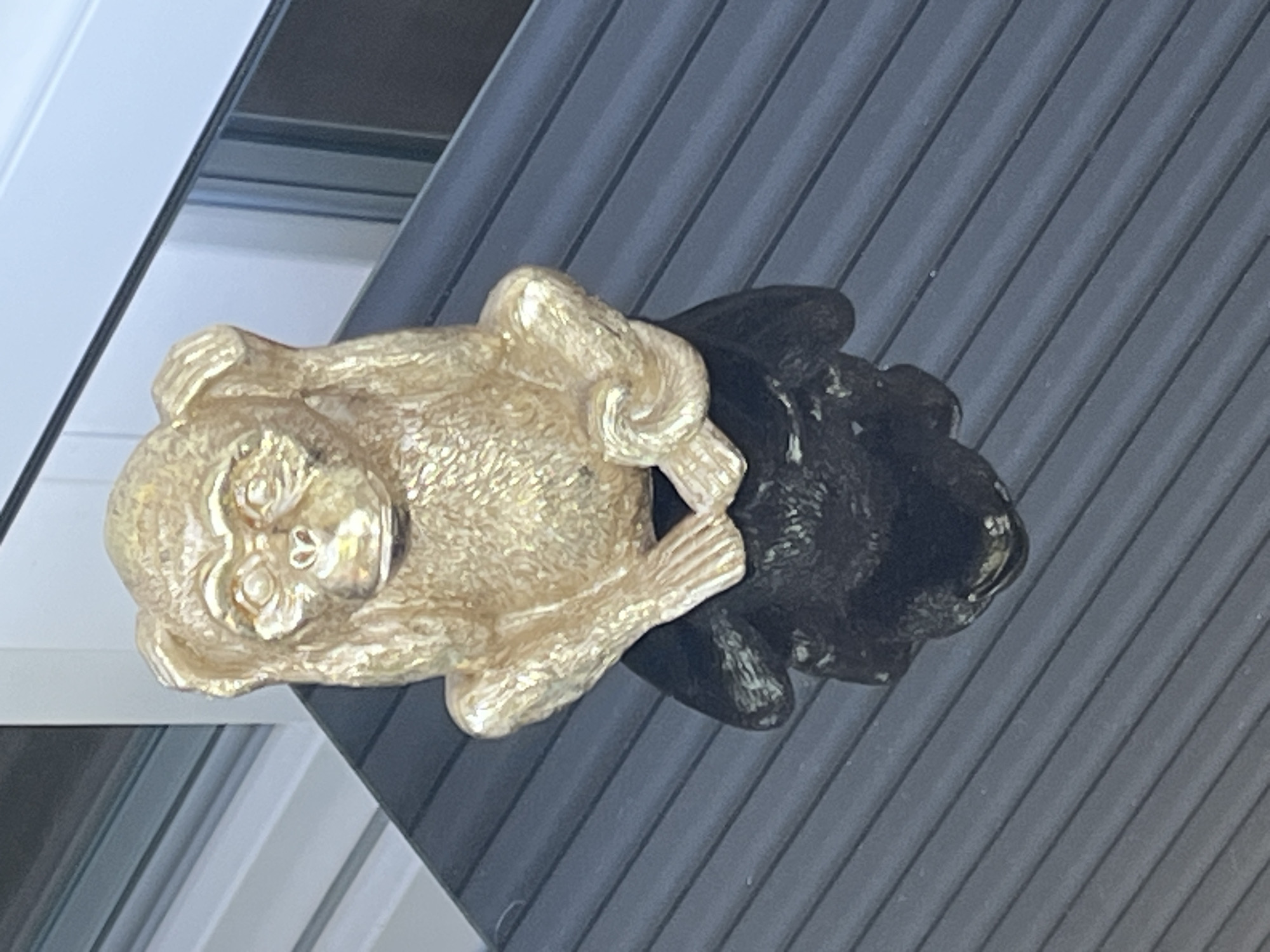 A golden statue of a monkey is sitting on a dark, ribbed surface, possibly a table or deck. The monkey is in a pose that suggests it is covering its ears. The background appears to be a reflection in a window, showing horizontal blinds and a white structure. The location is not specified in Garmisch-Partenkirchen, Germany.