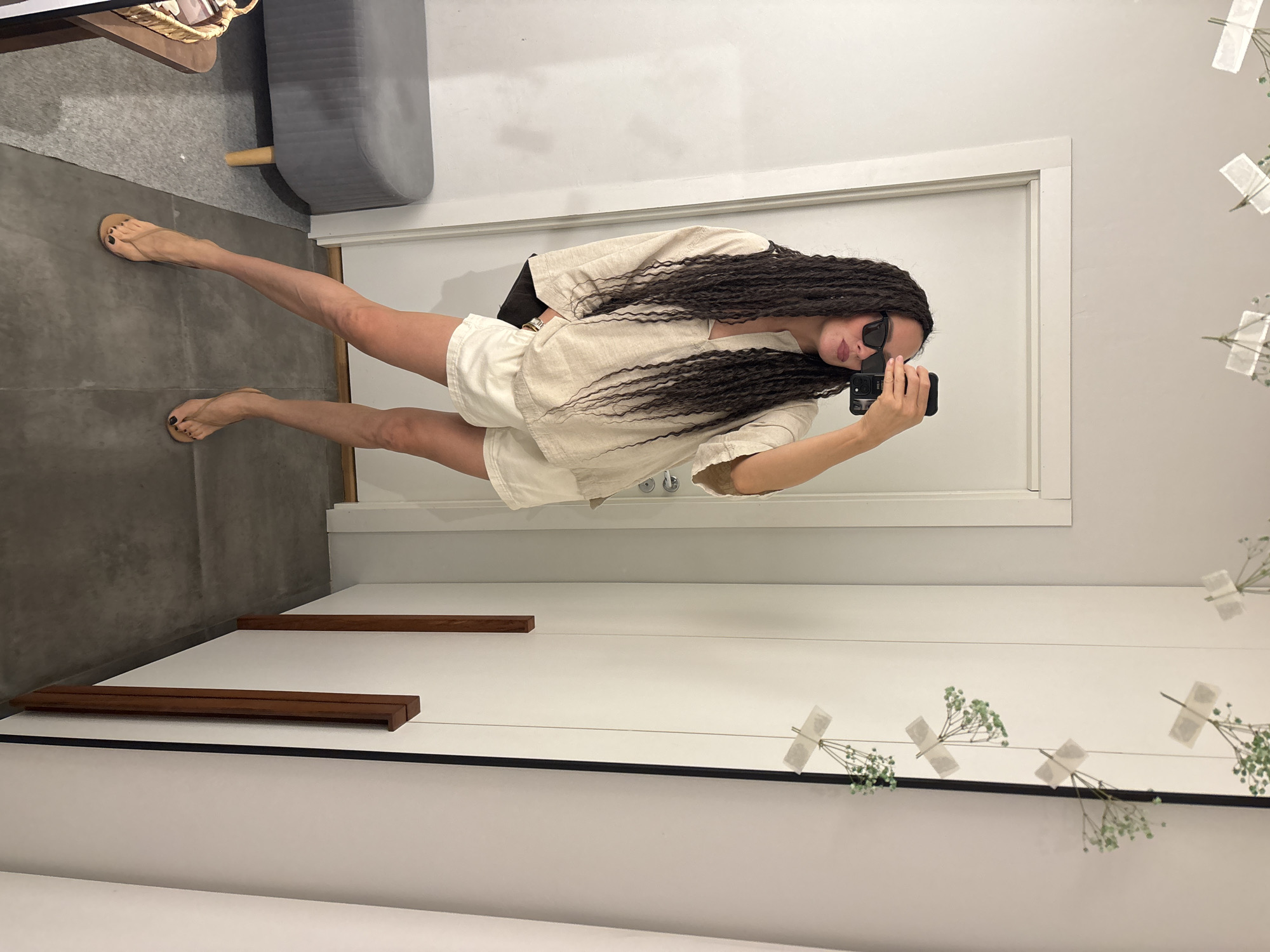 A full-body mirror selfie captures a woman in an indoor setting, likely a fitting room or retail space in Chaikovsky, Russia. The woman stands facing the mirror, holding a black smartphone with her right hand to take the photo. She has long, dark, braided hair or dreadlocks flowing down her shoulders and chest. Her eyes are obscured by black sunglasses, and her lips appear to have a subtle reddish tint.
She is dressed in a light-colored, possibly off-white or beige, two-piece outfit consisting of a relaxed-fit, short-sleeved V-neck top and matching shorts. On her feet, she wears light-colored flip-flops, and her toenails are painted a dark color, likely black. Her posture suggests a casual pose, with her left leg slightly bent.
The background visible in the mirror's reflection features plain white walls and a white door frame directly behind her. To the upper left, a grey upholstered stool with a light wooden leg is partially visible, along with a wicker basket situated on a surface above it. The floor is covered with dark grey tiles or concrete.
On the right side of the reflection, a decorative garland is affixed to the wall. It comprises small sprigs of green baby's breath (or similar foliage) taped to a thin string that stretches horizontally. Below this garland, two minimalist wooden rectangular shelf-like or display elements are mounted on the wall. The overall environment presents a clean and modern aesthetic. There is no visible text in the image.