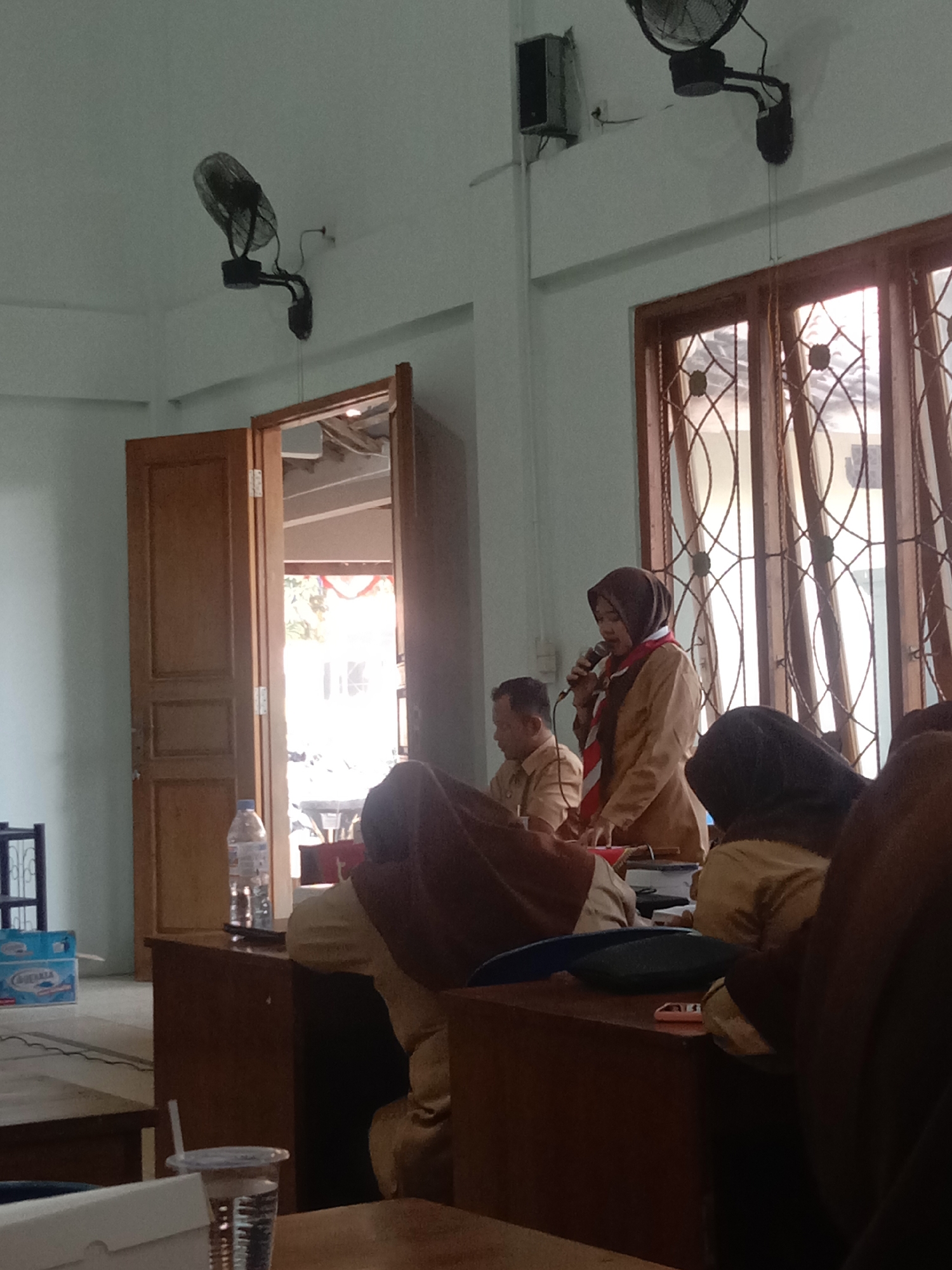 This is an indoor image, likely taken in Kalisari, Indonesia, showing a group of people, possibly students or attendees, in what appears to be a meeting or presentation. In the foreground, there are several individuals seated, some of whom are wearing brown uniforms with scarves. A woman in a brown hijab and a tan jacket is standing and speaking into a microphone, facing the seated individuals. Behind her, a window with intricate wooden lattice work allows natural light to illuminate the room. Two black oscillating fans are mounted on the ceiling, suggesting the need for ventilation. In the background, a wooden door is ajar, revealing a glimpse of an outdoor area. On a table in the foreground, there is a clear plastic cup with a straw and a clear bottle of water. A blue cardboard box is visible on the floor in the left background. The overall atmosphere seems to be one of a formal gathering or educational event.
