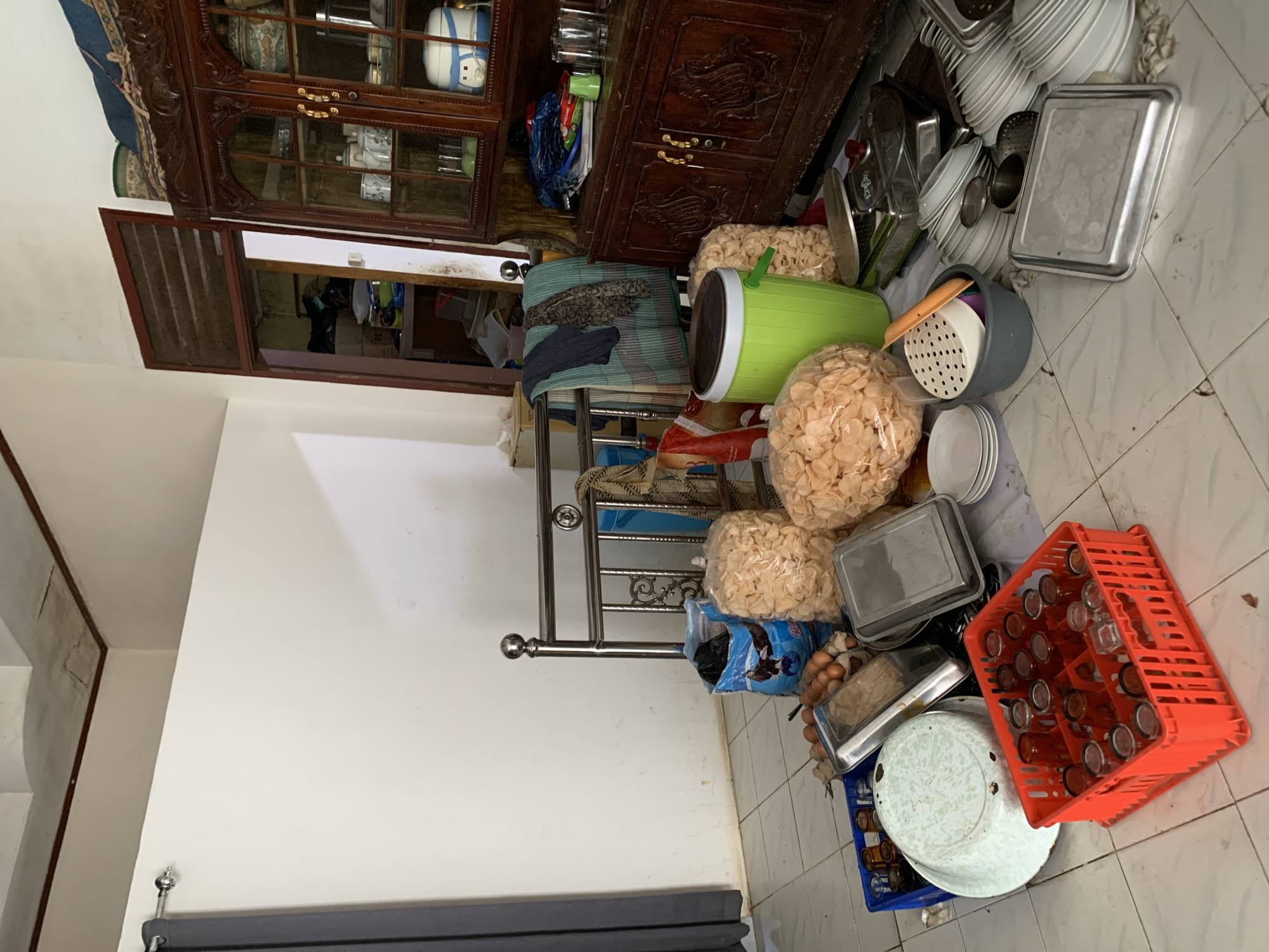 The image depicts a cluttered room in a residential setting in Uleepulo, Indonesia. The floor is tiled and appears somewhat grimy. In the foreground, an orange plastic crate contains numerous small amber glasses. To the left, a blue plastic crate holds more glasses. A large, white enamel basin with green speckles is also present. A metal tray holding a carton of brown eggs is visible. Several large plastic bags filled with light-colored, puffy snacks are scattered around. A bright green plastic bin with a lid stands near the center. To the right, a tall, ornate dark wood cabinet with glass doors displays various items, including glassware, a rice cooker, and decorative containers. Behind the cabinet, a doorway leads to another room that appears even more disorganized, with stacked boxes and miscellaneous items. A metal bed frame with decorative scrollwork stands against the white wall on the left, draped with blankets and clothing. A gray curtain hangs on the far left of the image. The overall impression is one of disarray and possibly preparation for an event or recent storage.