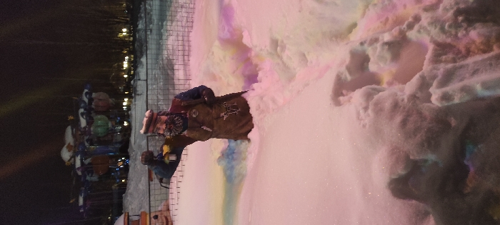 A festive scene unfolds at night. On the left, a carousel adorned with vibrant lights and figures stands near a dark fence. To the right, a person in winter attire is near a large pile of snow, bathed in an array of pink, purple, and blue lights. The snow appears soft and inviting, creating a whimsical atmosphere. Overhead, sparse trees are silhouetted against the night sky.