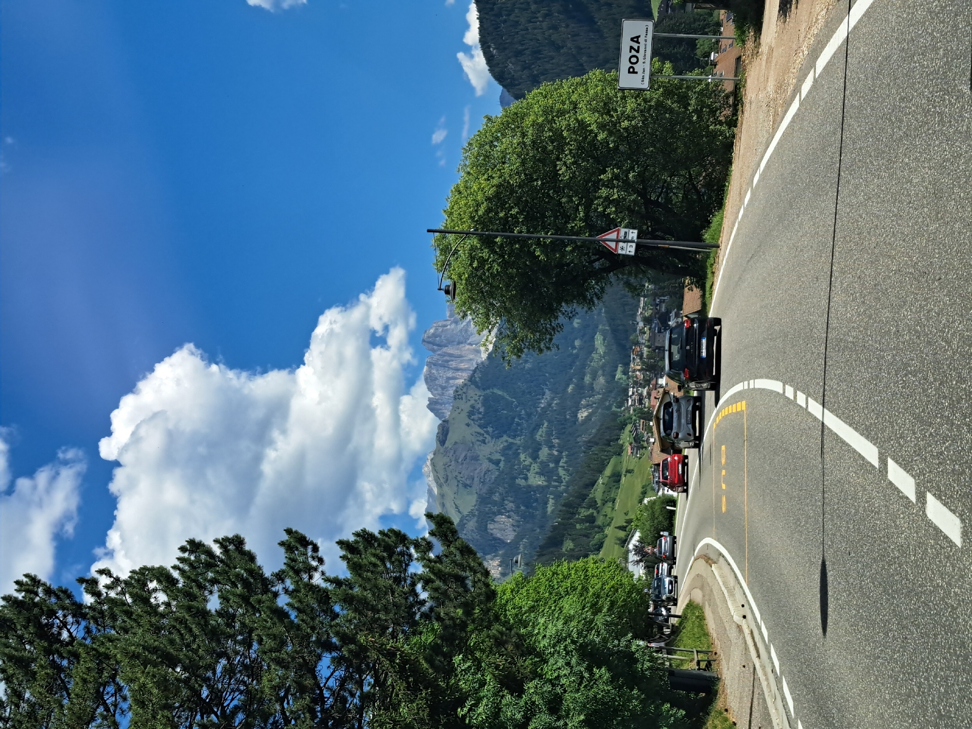A low-angle, eye-level shot captures a paved road winding through a mountainous, rural landscape under a bright blue sky with scattered white clouds. A large, dark green pine tree dominates the left side of the frame, its branches reaching upwards. On the right side, a large, lush green tree stands near a white sign that reads "POZA" in black capital letters, with smaller text below indicating "San Jan - S.Giovanni di Fassa." In the background, a town with red-roofed buildings is nestled on a green hillside, which rises to meet imposing, jagged grey mountains. Several cars are visible on the road, moving away from the viewer. One car, a black hatchback, is in the foreground, followed by a silver hatchback and a red hatchback. A yellow "BUS" marking is painted on the road surface. The road has solid white lines marking the edges and dashed white lines in the center. The environment appears to be a scenic route in a mountainous region, likely during daytime.