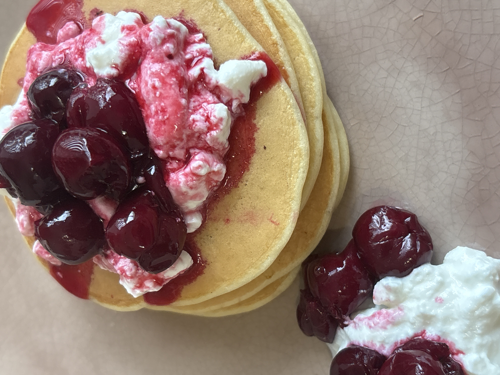 A close-up, high-angle shot presents a stack of pancakes on a pale, cracked plate. The pancakes are topped with a generous dollop of what appears to be cottage cheese, drizzled with a red sauce, and adorned with dark red cherries. A smaller portion of the same topping with cherries sits separately to the right of the pancake stack. The setting is likely a kitchen or dining area. The image contains no people or identifiable landmarks indicating it is in Saint Petersburg, Russia.