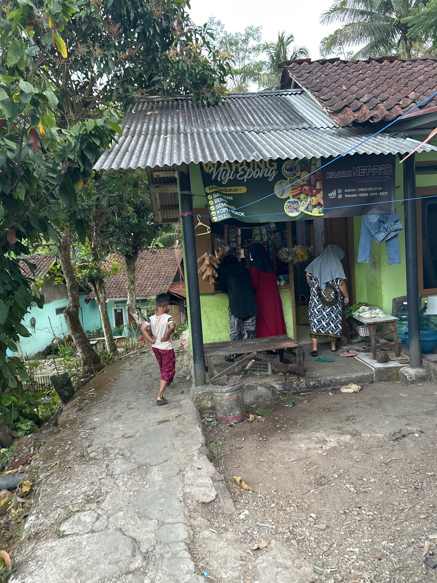 The image depicts a street-level view of a small, open-air shop in Cihaurbeuti, Indonesia. The shop has a corrugated metal roof and green painted walls, with a banner above the entrance that reads "Ngi Epong" and "HIDAYAH NET-PRO." The banner also lists services and products such as "SAYURAN" (vegetables), "BERAS" (rice), "Sembako" (staples), "KOPI" (coffee), and "DLL" (etc.), as well as contact information for an administrator and technician. A young boy, wearing a light-colored t-shirt and maroon shorts, stands on a concrete path to the left of the shop, facing away from the camera with his hands on his hips. Inside the shop, three individuals are visible. One person, wearing a red dress and headscarf, is looking at goods. Another person, wearing a dark jacket and patterned pants, is also looking at goods. A third person, wearing a grey headscarf and a black and white patterned dress, is standing with their back to the camera, facing into the shop. A wooden table to the right of the entrance holds some produce. In front of the shop, there are various items on the ground including shoes, a bucket, and debris. The environment appears to be rural or semi-rural, with vegetation and houses visible in the background. A palm tree is visible in the upper right corner.