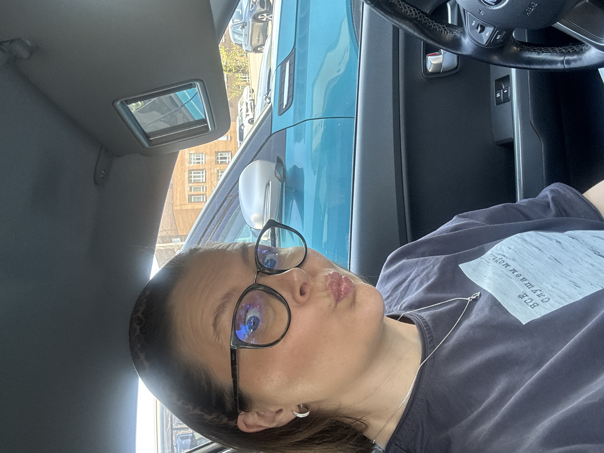 A woman is inside a car. She is wearing glasses, a gray t-shirt with a square image on it, and a silver necklace. The car's steering wheel and dashboard are visible. Through the car window, a building with multiple windows is visible. The exterior of a blue car is also visible. This appears to be Izhevsk, Russia.