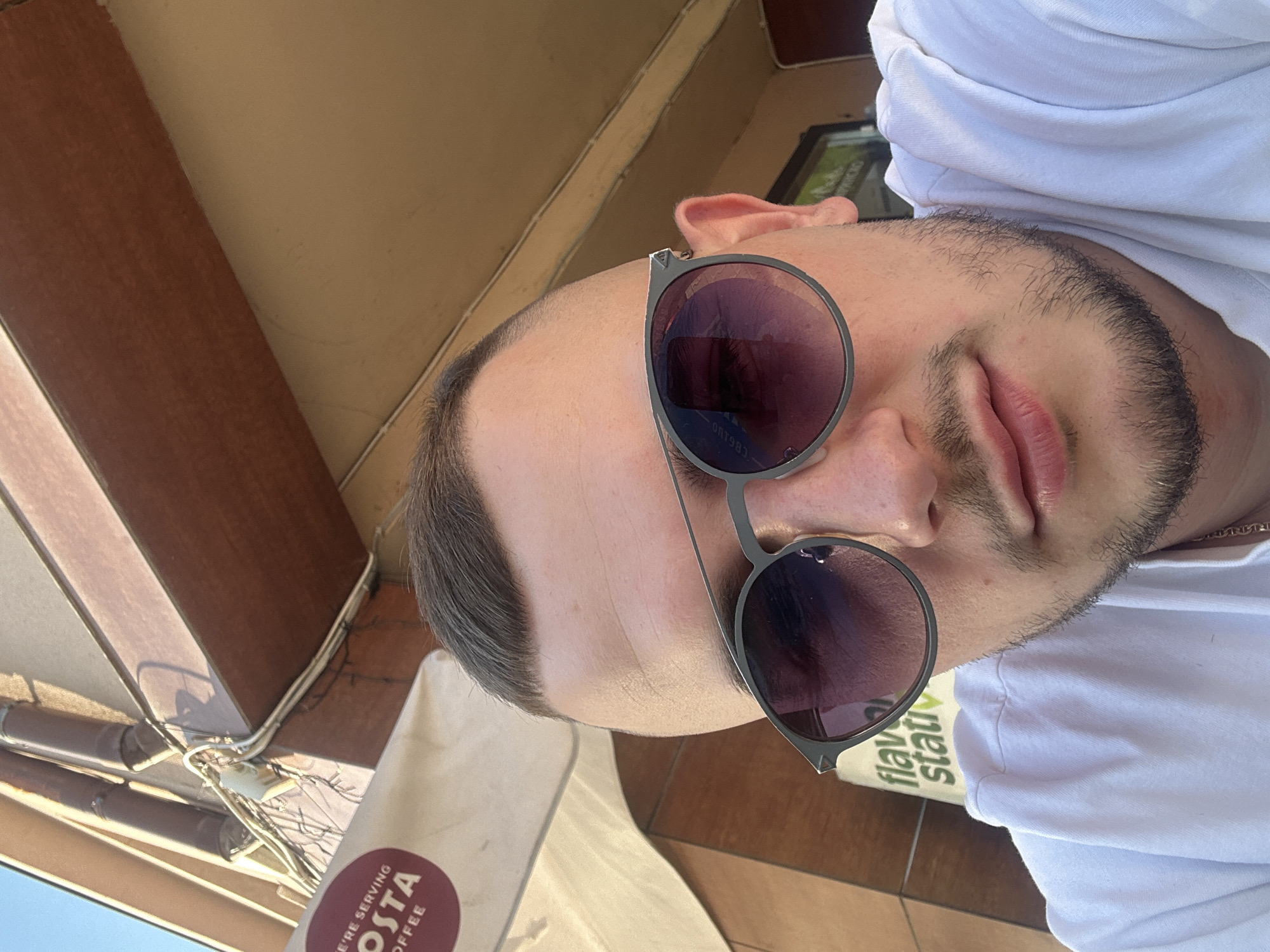 The image shows a close-up selfie of a man wearing sunglasses and a white shirt. The man has short hair and a beard. The backdrop includes parts of an outdoor setting, featuring elements of a building structure and the logo of Costa Coffee on a paper cup. The "flavour" text can be seen as well, indicating the context of a coffee shop. The location appears to be Plovdiv, Bulgaria, based on the potential presence of Costa Coffee, a chain that is present in this location.