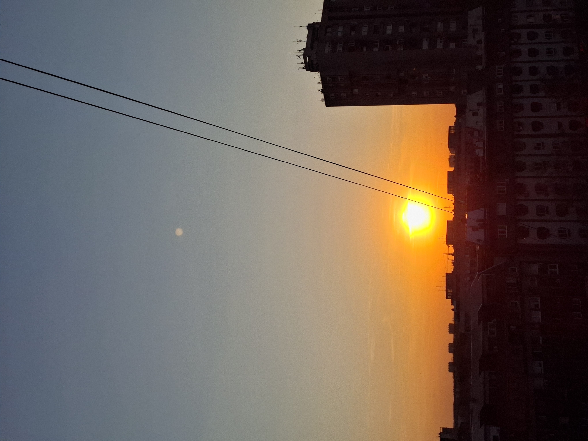 The image depicts a sunset in Novi Sad, Serbia. The sun is setting between buildings, creating an orange glow in the sky. Two electrical lines are visible in the sky. The image is rotated clockwise.