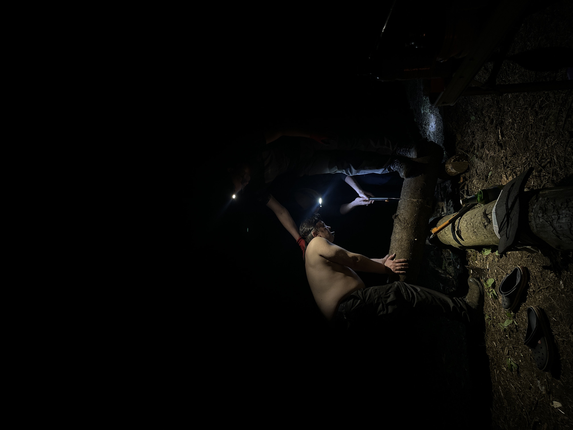 In a dark outdoor environment in Папярнянскі сельскі Савет, Belarus, two people are illuminated by headlamps. One shirtless man is seated, leaning against a tree trunk. Another person stands above, assisting with an object or activity involving the tree. An axe and footwear are visible on the ground nearby.