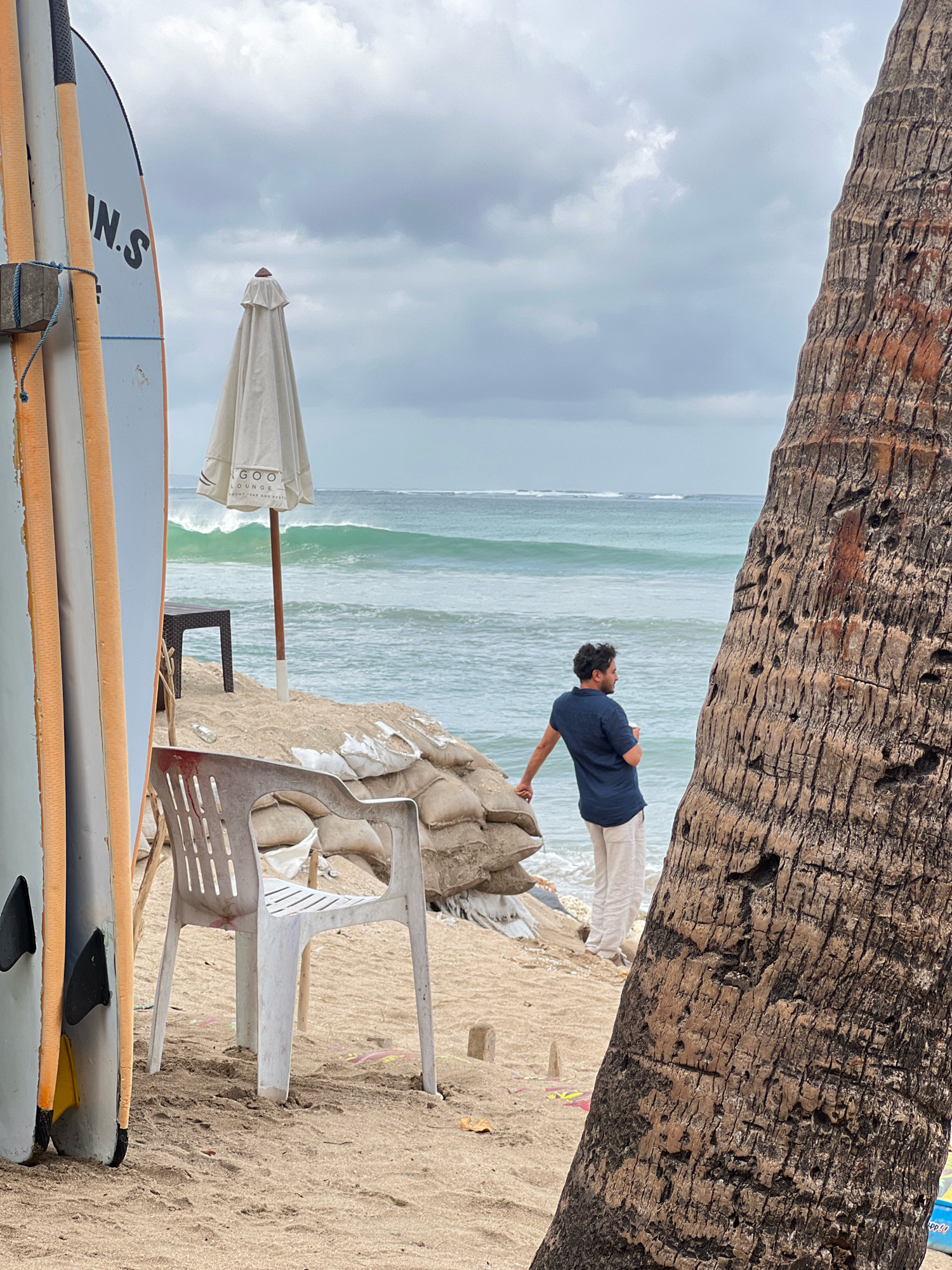 In Kuta, Indonesia, on a cloudy day, a man stands on a sandy beach near the ocean, looking out at the waves. He is wearing a dark blue short-sleeved shirt and light-colored pants. To his left, a large palm tree trunk dominates the foreground, its rough bark textured and weathered. In the background, the ocean exhibits moderate waves. A white beach umbrella, closed, stands on a slightly elevated section of sand, behind which are several sandbags, likely for flood control. In the left foreground, leaning against a wooden post, are several surfboards, their tips visible. A white plastic chair sits on the sand in front of the surfboards. The overall atmosphere is calm and somewhat overcast.
