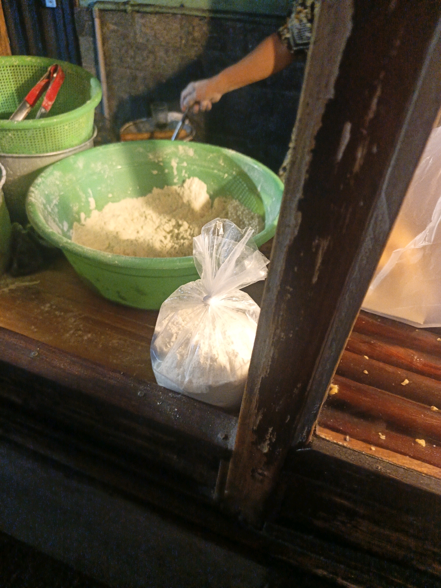 The image captures a close-up, low-angle view of a food preparation area, likely at a street stall in North Jakarta, Indonesia. In the foreground, a large green bowl is filled with a light-colored powdery substance, appearing to be flour or a similar ingredient. To the right of the bowl, a clear plastic bag containing a portion of the same powdery substance is tied with a string. A wooden structure, possibly part of the stall's counter or display, runs diagonally through the right side of the frame, showing some wear and tear with visible smudges and debris. In the background, a portion of a green colander holding red-handled tongs is visible, suggesting ongoing food preparation. A person's arm and hand are seen in the upper right portion of the frame, engaged in some activity that is not entirely clear but involves interaction with a metal utensil over a pan, hinting at cooking or frying. The environment appears to be outdoors or in an open-air market setting, with dim lighting contributing to a somewhat rustic and busy atmosphere.