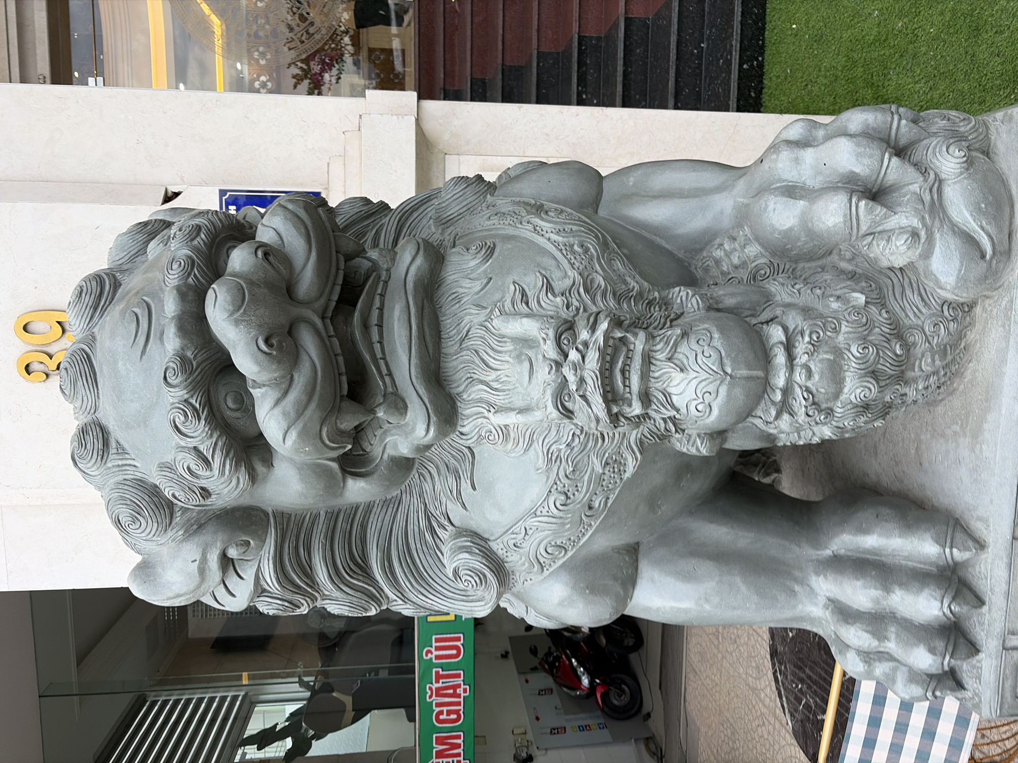 A close-up, low-angle shot captures a large, gray stone Foo dog statue positioned in front of a building in Da Nang, Vietnam. The statue is highly detailed, featuring a fierce expression with bared teeth and intricately carved mane and fur. It is adorned with a smaller dragon-like creature on its chest. Behind the statue, a glass-fronted building is visible, with reflections of the interior and exterior. To the left, a green sign with Vietnamese text is partially visible. To the right, a set of dark stairs with red risers leads up the side of the building, with a patch of green artificial grass in the foreground. The overall atmosphere is one of cultural symbolism and architectural presence.