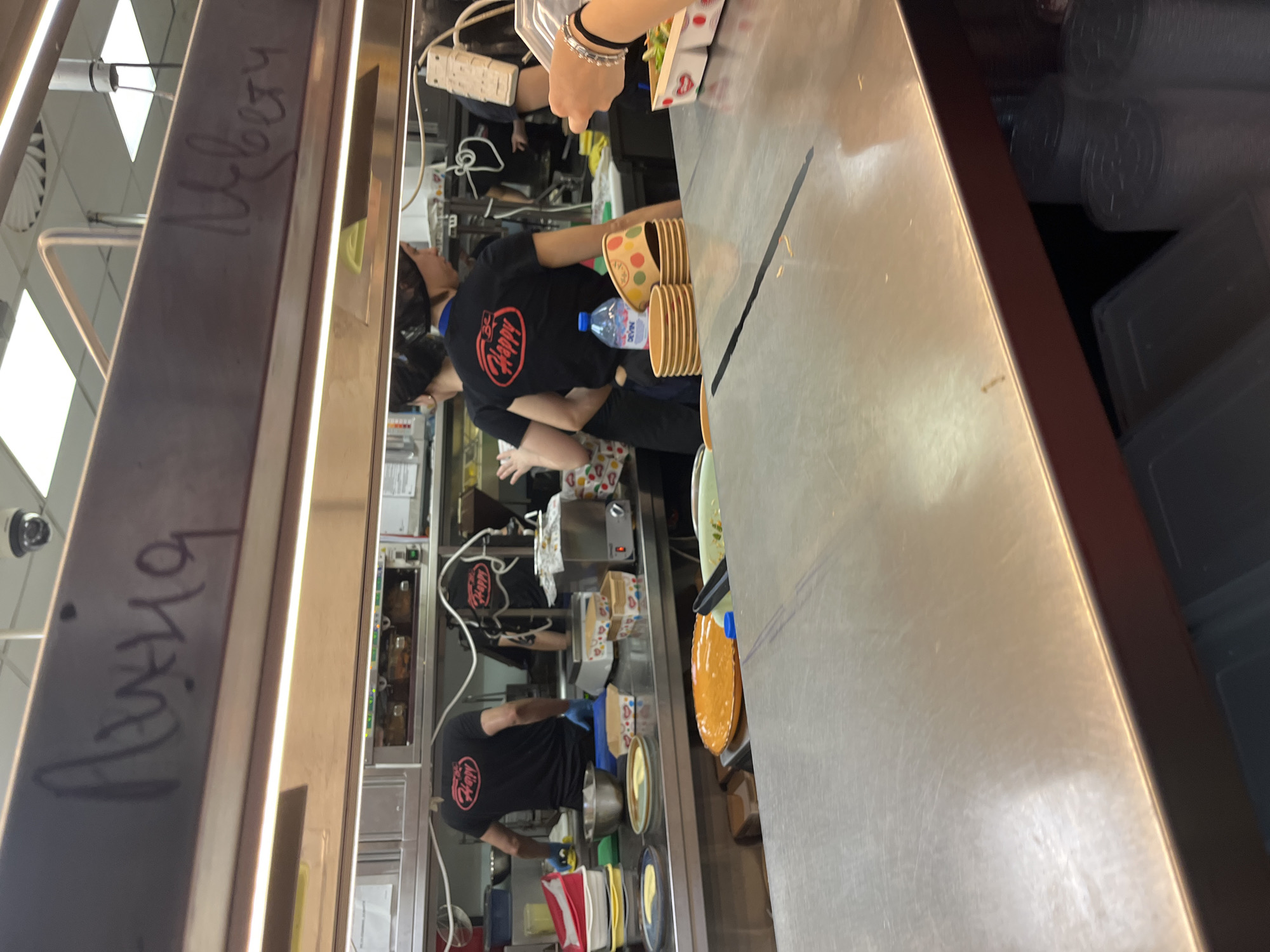 Here's a detailed description of the image:
**Location and Context:** The image appears to be taken inside a kitchen or food preparation area of a restaurant, possibly in Varna, Bulgaria. This is suggested by the Cyrillic script visible on the metallic structure in the foreground, which might indicate a local setting.
**People:** There are multiple individuals visible in the image. They seem to be restaurant staff, as they are wearing black t-shirts with a logo (likely the restaurant's logo) and appear to be engaged in food preparation tasks.
**Objects:**
* Several disposable cups are stacked and near what looks to be a water bottle.
* Various plates and dishes are visible, suggesting different stages of food preparation.
* Food preparation equipment and utensils are present, indicating a working kitchen.
* A large stainless steel countertop dominates the foreground.
* Plastic containers are stacked on the side.
**Actions and Interactions:** The people are actively working on food preparation. This is implied by the presence of food items, plates, and utensils, as well as the fact that they are wearing work attire.
**Environment:** The environment is a functional kitchen space with stainless steel surfaces, presumably for hygiene. There are also storage areas for food containers.