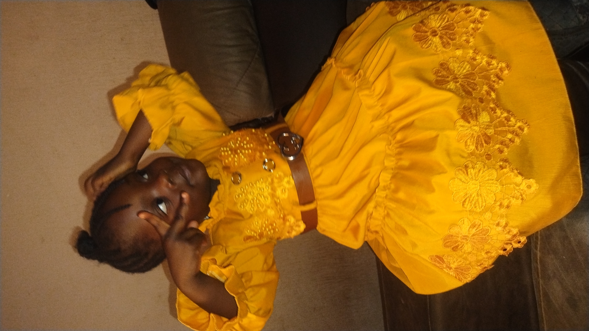 A young child lies on a couch or cushioned surface. The child wears a bright yellow dress adorned with floral embroidery along the hem of the skirt. A brown belt with a silver buckle cinches the waist of the dress. The child has dark skin and short braided hair pulled into a small bun. The child is looking up, holding two fingers near their face. The background includes a beige wall or floor and the brown leather of the couch. The location is identified as Jos, Nigeria.