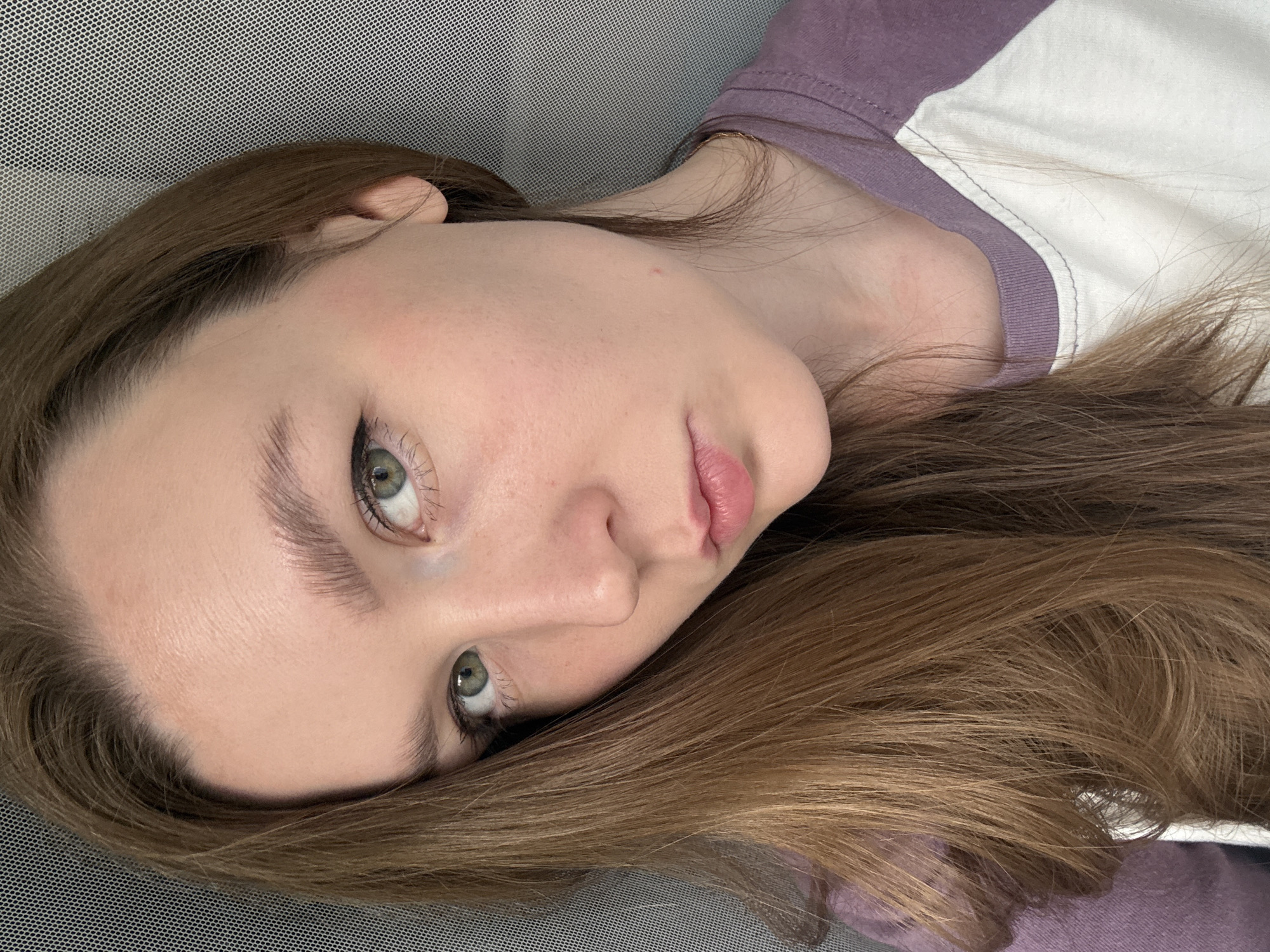 A close-up, high-angle portrait features a young woman lying down, looking directly at the camera. Her head is tilted, and her long, straight, light brown hair cascades around her. She appears to be indoors, resting against a grey, perforated mesh surface, possibly a headboard, cushion, or chair back.
The woman has fair skin, with visible pores and a few minor, subtle skin imperfections, such as a small red spot on her right cheek and some faint texture. Her eyes are green or hazel, accentuated with black eyeliner and mascara on her lashes. Her eyebrows are full and appear naturally shaped. Her lips are a natural pink, slightly parted, with a soft, relaxed expression. She wears a casual t-shirt that is purple on the shoulder and upper arm, transitioning to white on the main body, indicating a raglan style.
The overall mood is calm and introspective. There is no specific activity depicted other than her looking at the camera. The lighting is soft and even, suggesting an indoor setting without harsh direct light, so the time of day and weather are not discernible. No visible text is present in the image. While provided with a location context of Izhevsk, Russia, there are no visual cues within the image itself to confirm this geographical location.