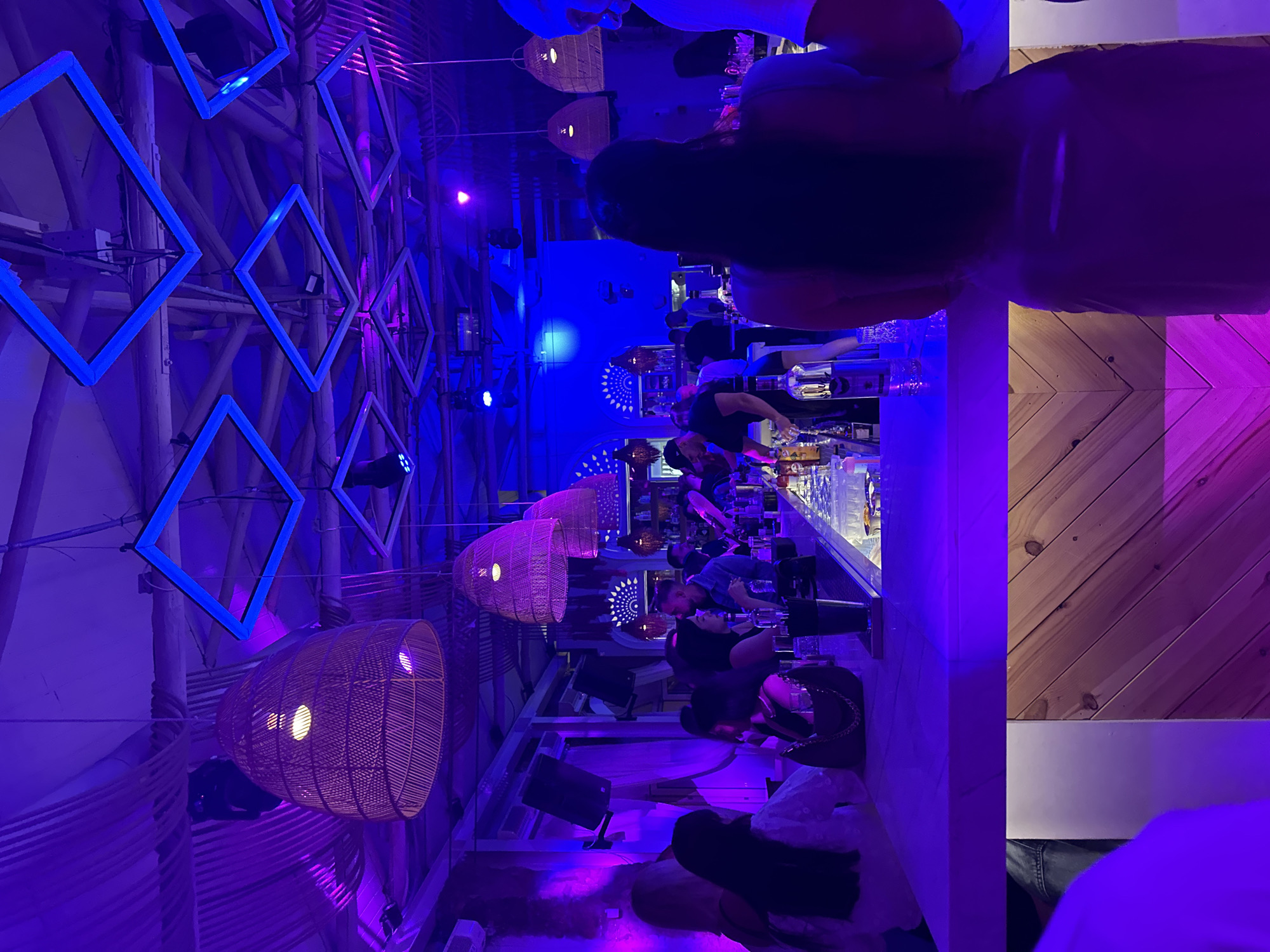 A dimly lit indoor bar scene in Pallini Municipal Unit, Greece, features hanging wicker lamps, square blue light fixtures, and a long bar with patrons. People are seated and standing at the bar, some interacting with the bartender. Wooden panels adorn the wall. The lighting is predominantly blue and purple.