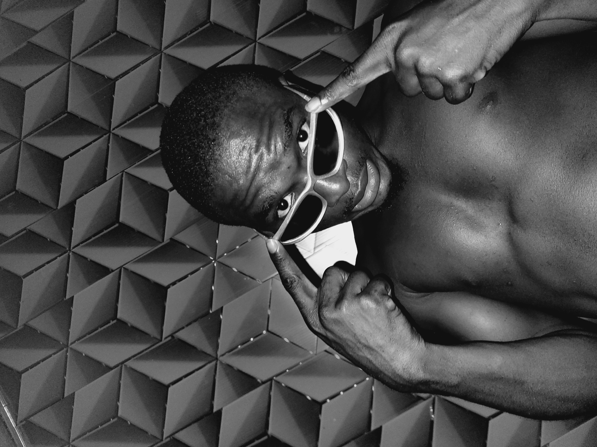 A close-up, black and white, medium shot shows a Black male with short hair, shirtless, wearing sunglasses. The man is looking at the camera, and his hands are on the sides of the sunglasses, pointing towards his face. The background is a geometric patterned wall. The image appears to be taken indoors.