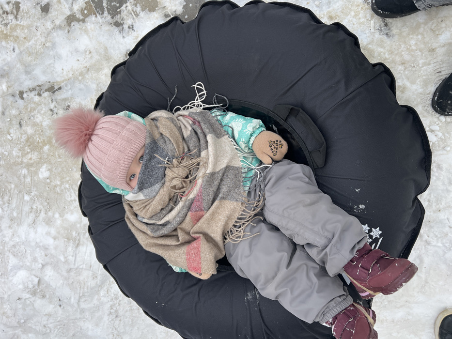 An infant or toddler, heavily bundled in winter attire, is observed sitting upright within a black inflatable snow tube on snow-covered ground in Primorka, Russia. The child wears a light pink ribbed knit beanie with a large pink pom-pom, a multi-colored plaid scarf (tan, brown, grey, red) wrapped around the neck and lower face, obscuring the mouth and chin, and a light blue patterned winter jacket. Grey snow pants and dark red boots with Velcro fasteners cover the lower extremities. Tan mittens with dark grip dots are visible on the hands. The child's blue eyes are visible above the scarf. The immediate environment consists of patchy snow and ice. The toes of dark footwear, presumably belonging to an adult, are visible in the lower left and right corners of the frame, suggesting proximity to another individual.