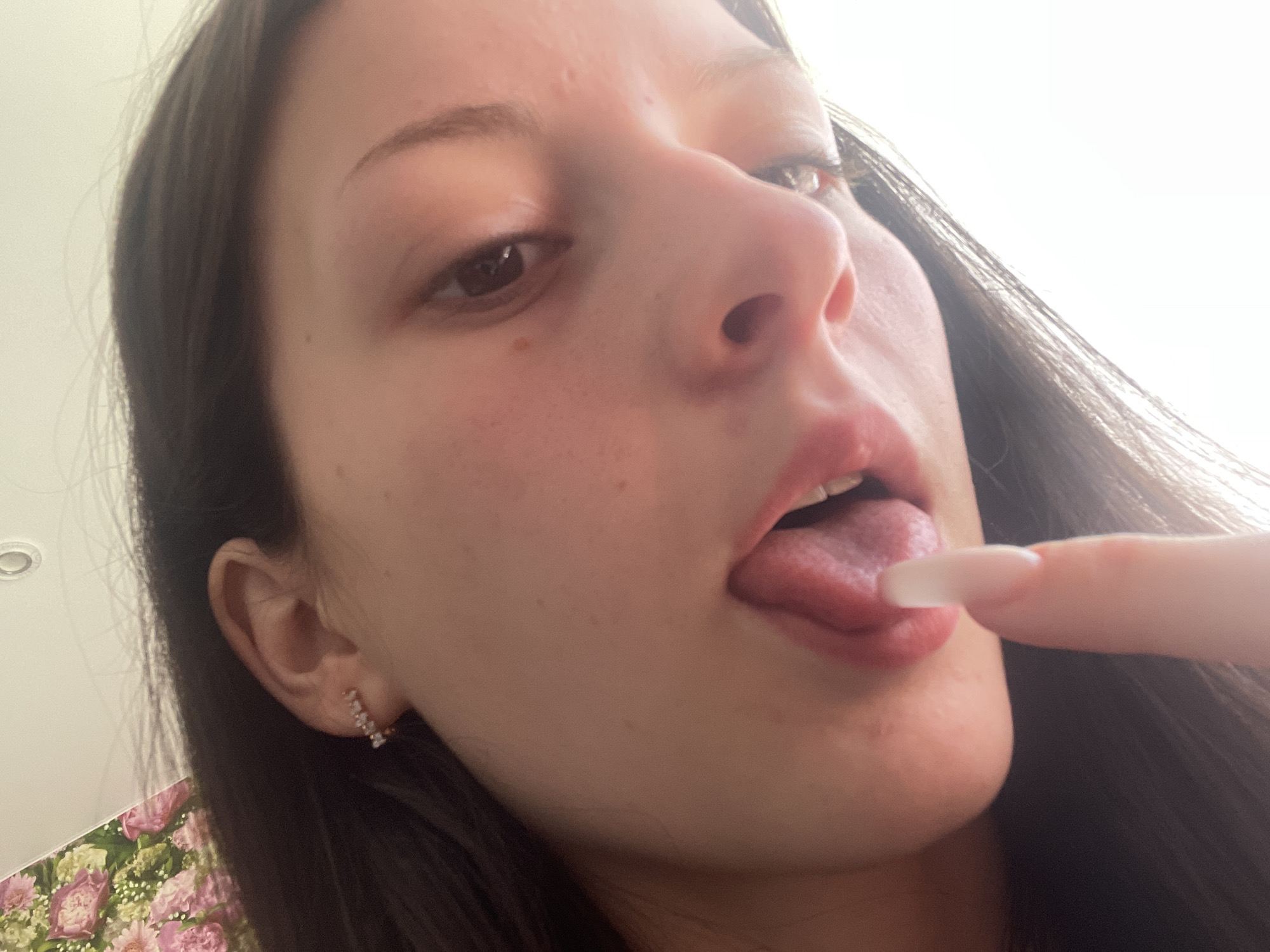 A close-up, low-angle shot captures a young woman's face, primarily focusing on her mouth and nose, in what appears to be an indoor setting in Medwedewo, Russia. The woman is engaged in a playful or expressive act, sticking out her tongue while lightly touching it with the tip of her index finger, which features a long, light-colored manicured nail.
Her dark hair frames the right side of her face, and her left ear, adorned with a small, decorative earring featuring multiple small gems, is visible on the left. Her eyes are open and directed slightly upwards and to the side. The lighting is bright, almost overexposed in parts of the background, particularly on the upper right, suggesting a strong light source from above or behind her.
Visible details include a small portion of a white ceiling light fixture in the upper left corner and a soft floral pattern (pink and green hues on a light background) on what appears to be fabric, possibly clothing or bedding, in the lower left quadrant of the image. The overall impression is one of casual, intimate interaction or a playful pose. No text is visible in the image.