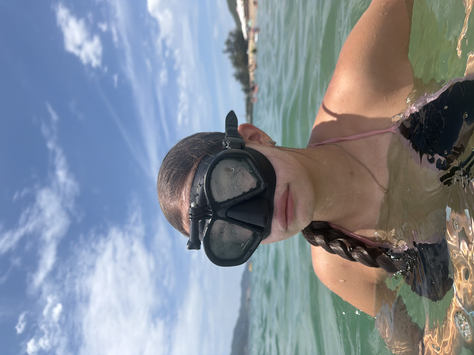 A young person wearing a black diving mask with a GoPro attached to it is in the ocean, likely near a beach in Nha Trang, Vietnam. The mask covers their eyes and nose, with only their mouth and chin visible. Their hair is styled in a dark braid that falls over their shoulder. They are wearing a black and pink bikini top. The water is clear and green, with ripples on the surface reflecting the bright blue sky and scattered white clouds. In the background, a coastline with trees and mountains is visible, along with a beach crowded with people and a few white umbrellas. The overall scene depicts a person enjoying a day at the beach and in the ocean.
