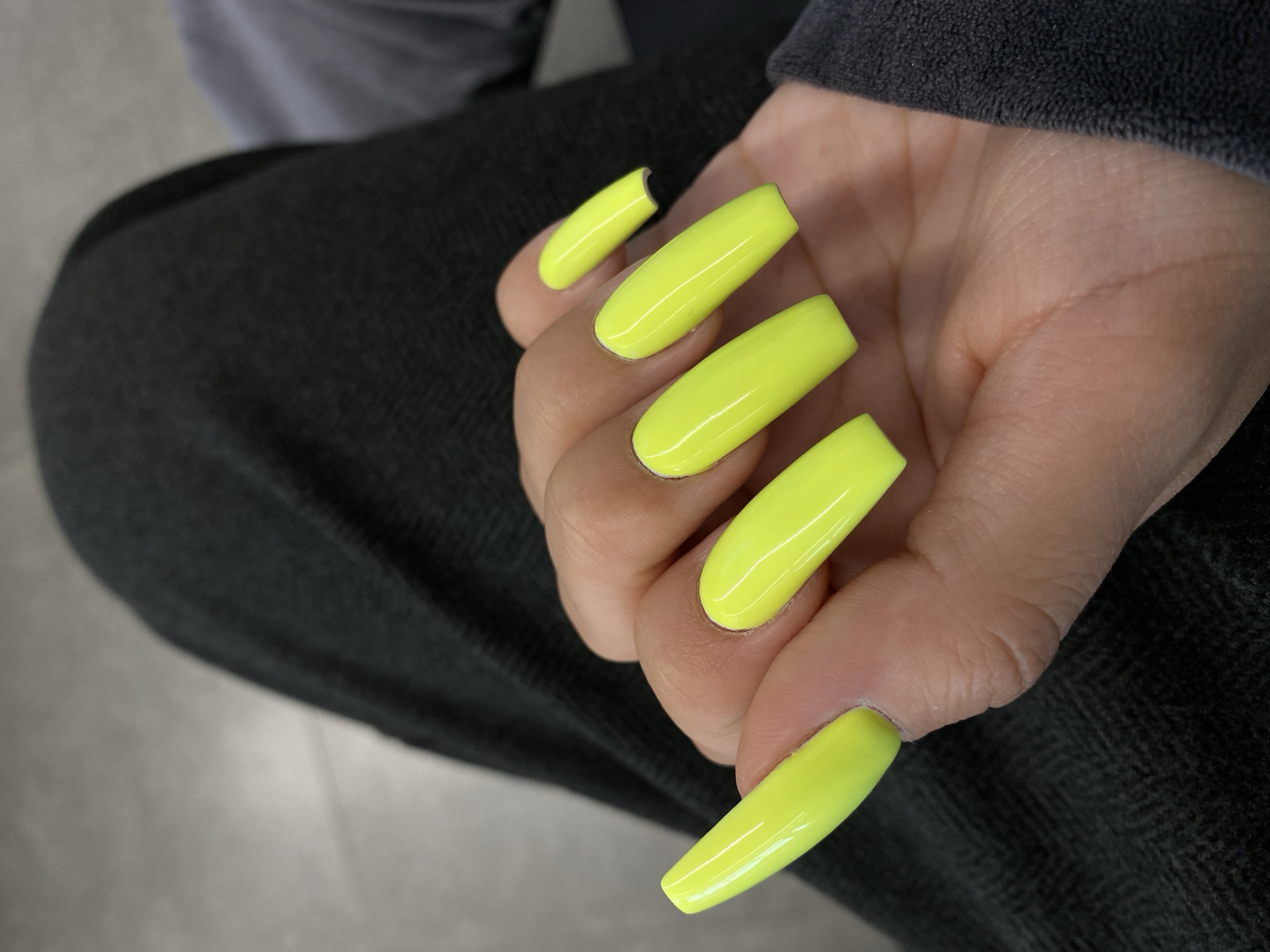 The image shows a close-up of a person's hand with long, bright neon yellow coffin-shaped nails. The hand is resting on dark gray fabric, possibly pants or a jacket, which has a subtle herringbone pattern. The background is a blurry gray surface, likely a floor or wall. The lighting is bright, highlighting the artificial gloss of the nails. The location is specified as Novoye Martyanovo, Russia.