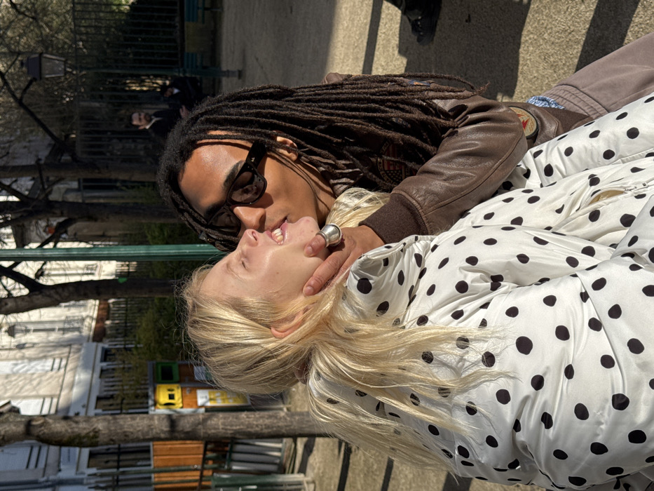 The image depicts two individuals engaged in an affectionate interaction within a Parisian public park under bright sunlight. A person with long, wavy blonde hair, wearing a white puffer jacket with black polka dots, is positioned on the left, their head tilted back with eyes closed and mouth slightly open, exhibiting an expression of pleasure. A minor blemish is visible near the left corner of their mouth. On the right, a person with dark dreadlocks, wearing black sunglasses and a brown leather jacket over a blue plaid shirt, has their left hand cupping the blonde individual's jaw. A prominent silver ring is on their left ring finger. This individual leans in, their lips making contact with the blonde person's lower cheek or the corner of their mouth. The background reveals classic Parisian Haussmannian architecture, specifically light-colored stone buildings with dark windows and balconies. The park environment includes bare deciduous trees, a dark green metal fence, and a traditional dark streetlamp. The ground is a light-colored gravel path. Two additional, indistinct figures are visible in the distant background, one standing and one seated on a bench. The setting is a public square or park in Paris, France.