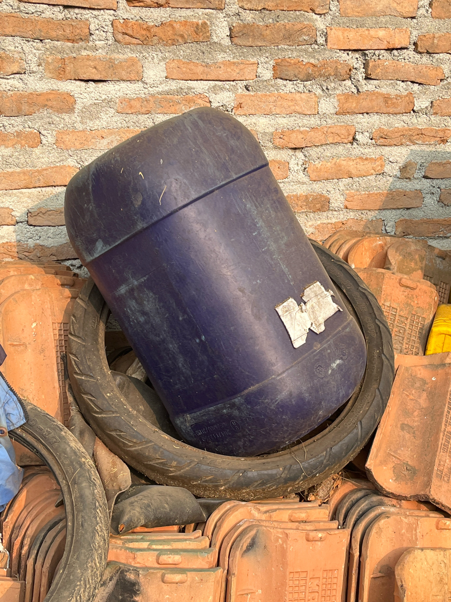 A pile of terracotta roof tiles is stacked in front of a rough brick wall in Kebumen, Indonesia. A large, dark blue plastic drum is tilted on top of the tiles, supported by a motorcycle tire. The drum has a white sticker partially attached to its side, and some dirt and debris are visible on its surface. The motorcycle tire is worn and appears to have some writing on its sidewall. Another motorcycle tire is visible to the left, partially obscured by a blue garment. A yellow container is also visible in the background, to the right of the drum. The scene is illuminated by natural light, casting shadows on the ground and wall.