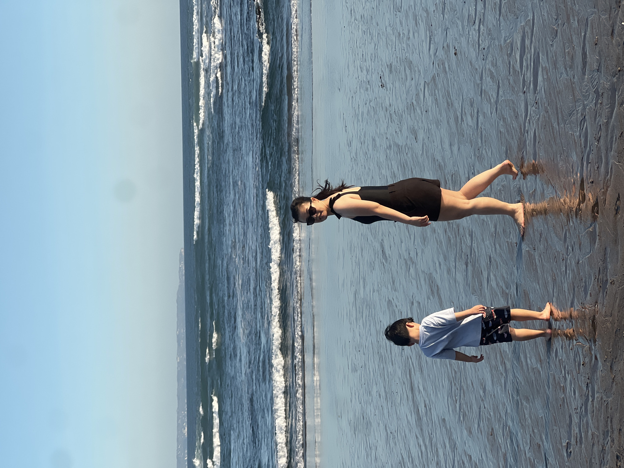 This is a vertical, outdoor photograph taken at eye-level on a beach in North Kuta, Indonesia. The scene depicts two people: a woman and a young boy, standing on wet sand. The woman, on the left, wears a black dress and sunglasses, looking towards the camera with a slight smile. She is positioned closer to the water's edge, with gentle waves lapping at the shore. The boy, on the right, wears a light blue t-shirt and patterned shorts, looking down and away from the camera. His stance suggests he might be contemplating something or observing the sand.
The setting is a beach with a vast expanse of wet sand, reflecting the sunlight. The ocean is visible in the background, with white-capped waves breaking on the shore. The sky is a clear, bright blue, indicating a sunny day. The lighting suggests it is daytime, likely late morning or early afternoon. The overall mood of the image is relaxed and peaceful, capturing a moment of leisure and enjoyment. There are no visible text elements in the image. The wet sand creates a textured, almost rippled surface that adds visual interest to the foreground.