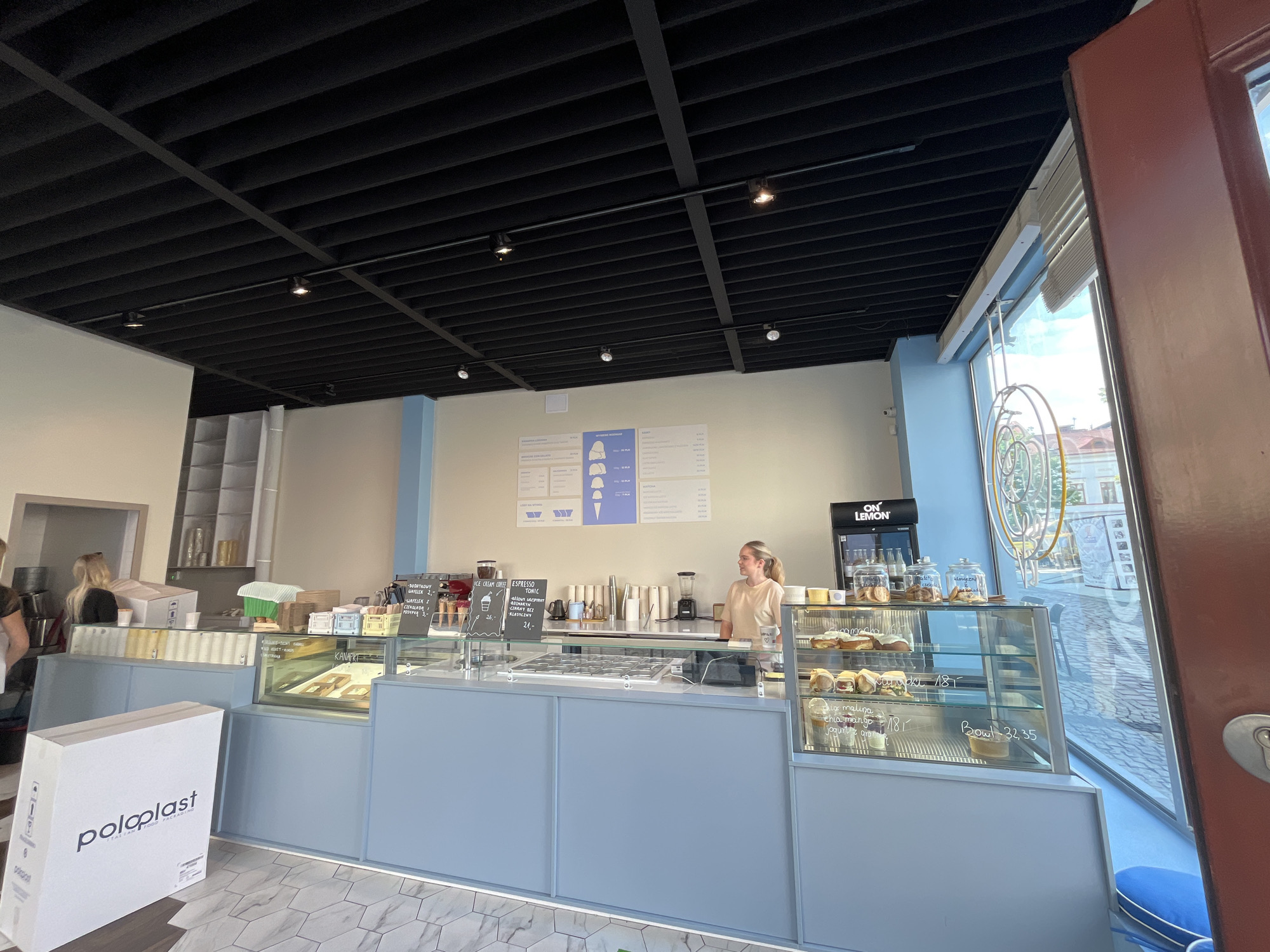The image shows the interior of a cafe. The cafe has a light blue counter with glass display cases filled with pastries and jars of cookies. A young woman with blonde hair tied in a ponytail is standing behind the counter, wearing a light peach-colored shirt. She is looking to her right. To the left of the counter, there is a large white box with the logo "poloplast" printed on it. The ceiling is black with a grid of lights. A large window on the right side of the image looks out onto a street with buildings and trees. The floor has a hexagonal tile pattern. There are menus on the wall behind the counter displaying prices for ice cream, coffee, and bowls.