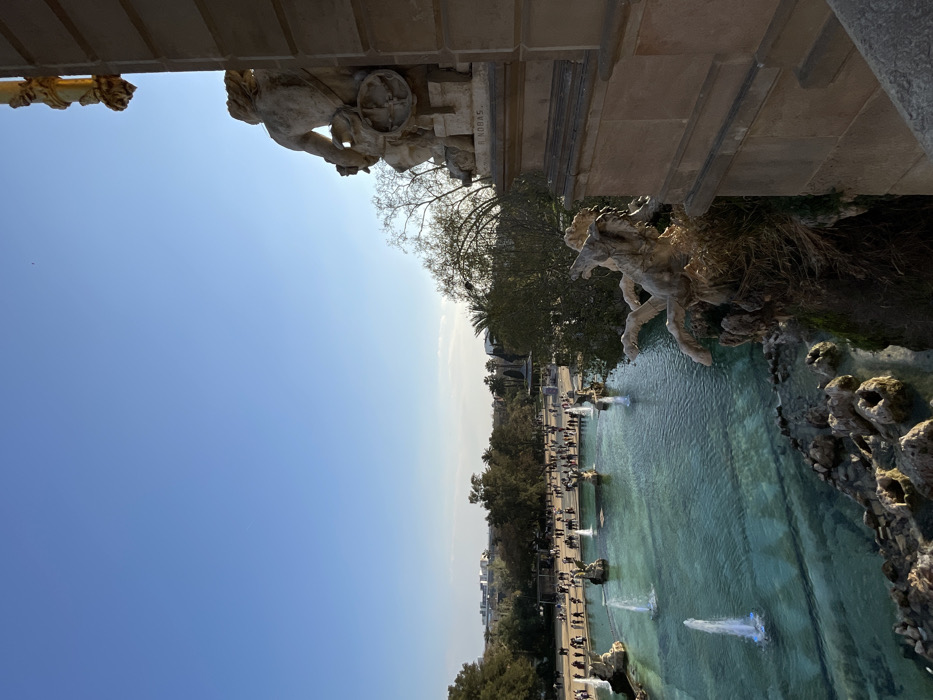 The image displays an elevated perspective of the Cascada Monumental located within Parc de la Ciutadella, Barcelona, Spain. The sky is clear blue with minimal cloud cover, indicating daytime.
In the immediate right foreground, a large, light-colored stone statue of a muscular male figure is visible from the rear, integrated into a multi-tiered architectural structure. The name "R. NOBAS" is carved into the base of this statue. Directly beneath it, another intricate stone sculpture depicts a dynamic mythological creature, possibly a sea horse, emerging from a rocky, vegetated cascade that feeds into the main water basin.
The central midground features a large, turquoise-blue water basin with multiple active water jets producing fountains. Several darker-colored statues resembling winged creatures are situated within this basin. Beyond the fountain, a wide expanse of the public park is visible, densely populated with numerous individuals. These people are observed walking and congregating on paths and open grassy areas throughout the park. The park environment includes a variety of mature trees, both deciduous and evergreen, including palm trees. Distant urban buildings are discernible on the horizon line behind the park's tree canopy. The entire scene is brightly illuminated by direct sunlight, casting distinct shadows.