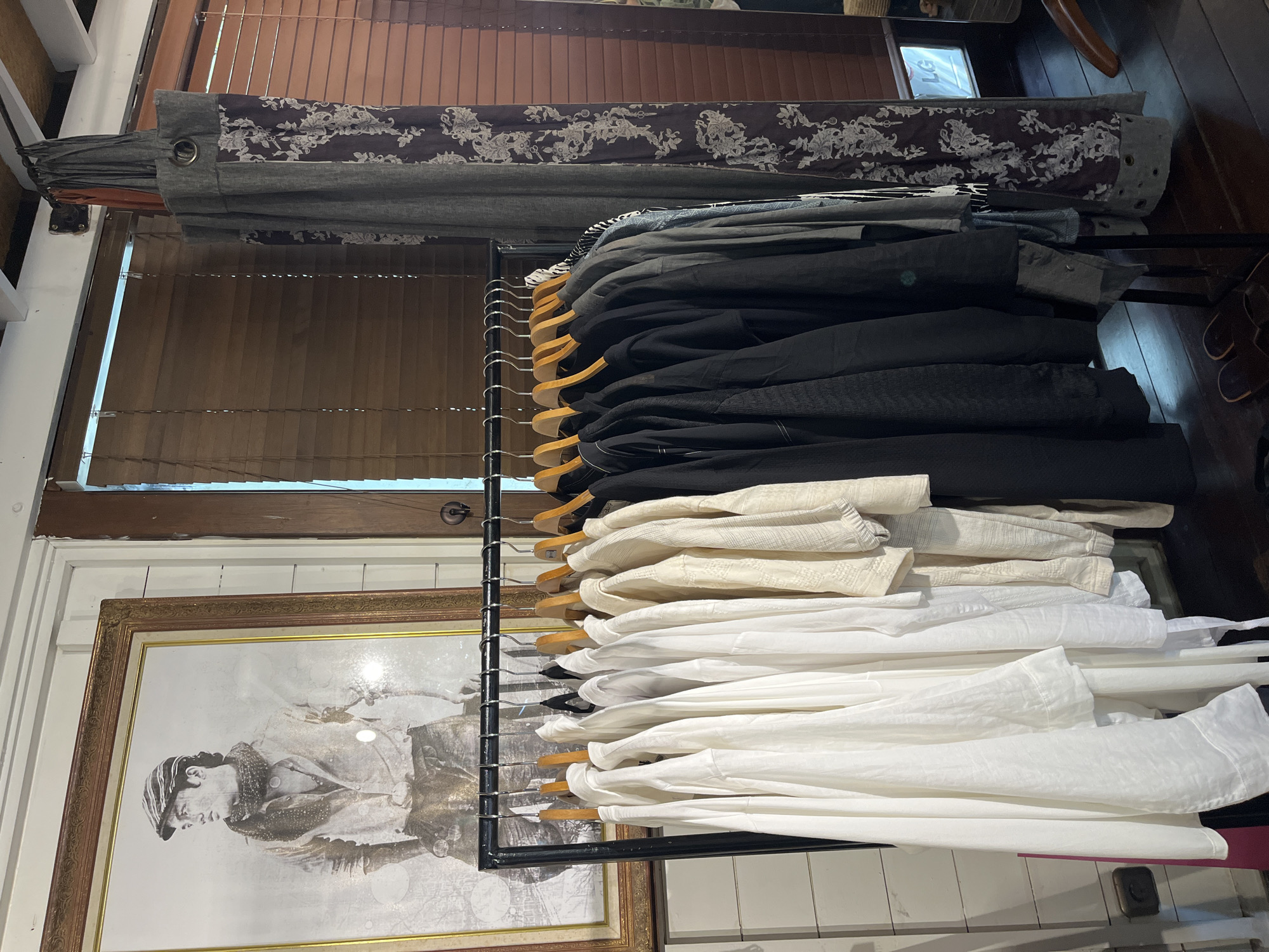 The image depicts a clothing rack filled with various garments, predominantly white and black shirts, hanging on wooden hangers in what appears to be a retail setting. Behind the rack, a large framed black and white photograph of a man wearing a flat cap and scarf is visible. The background features wooden blinds and a patterned curtain. The scene is well-lit, suggesting an indoor environment. The location is identified as North Kuta, Indonesia.