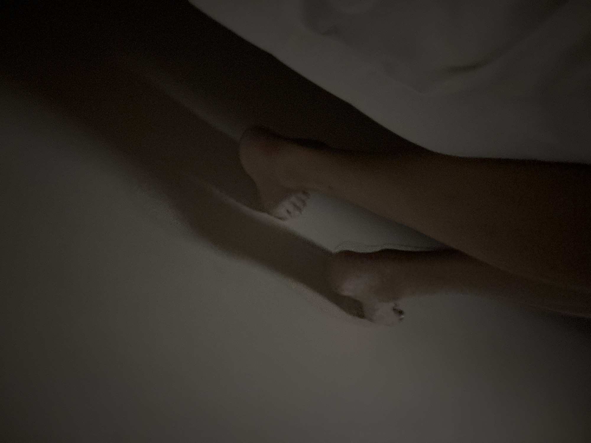The image captures a dimly lit, intimate scene featuring a person's lower legs and feet, partially covered by a light-colored sheet or blanket. The lighting is very low, casting soft shadows and indicating either nighttime or a very dark room. One foot is more clearly visible towards the center-right, with distinct toes, while the other leg and foot are largely obscured by the fabric and shadows in the foreground. The visible skin tone is light. The person appears to be resting or sleeping in a bed. No specific location cues, weather details, or visible text are present within the image. The overall impression is one of peaceful rest.