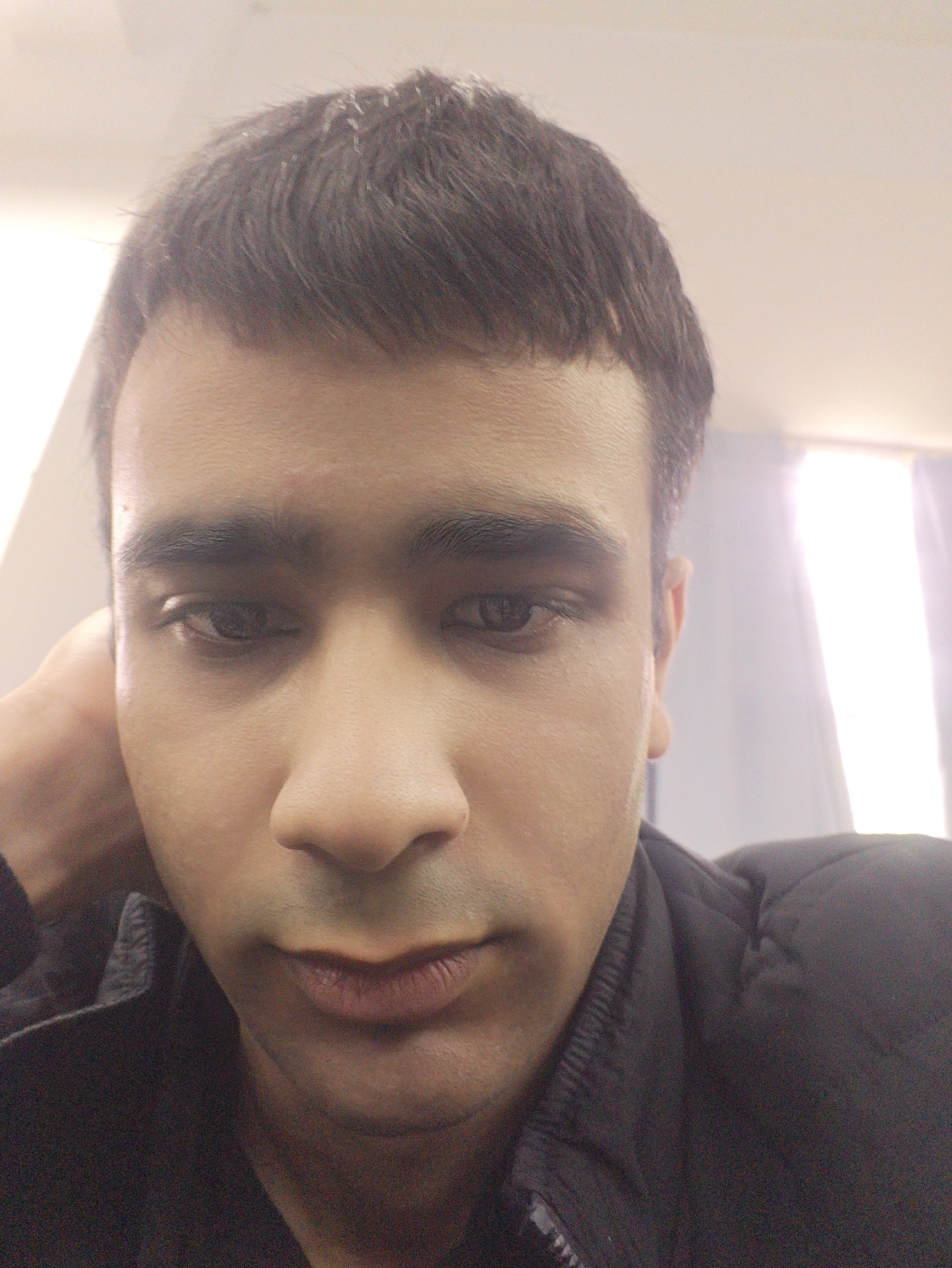 A close-up selfie of a man with dark hair and complexion, wearing a dark jacket. The background is indistinct, with some light from a window. The image does not provide specific evidence to confirm the location as Chongqing High-tech Zone, China. There are no actions or interactions taking place in the image.