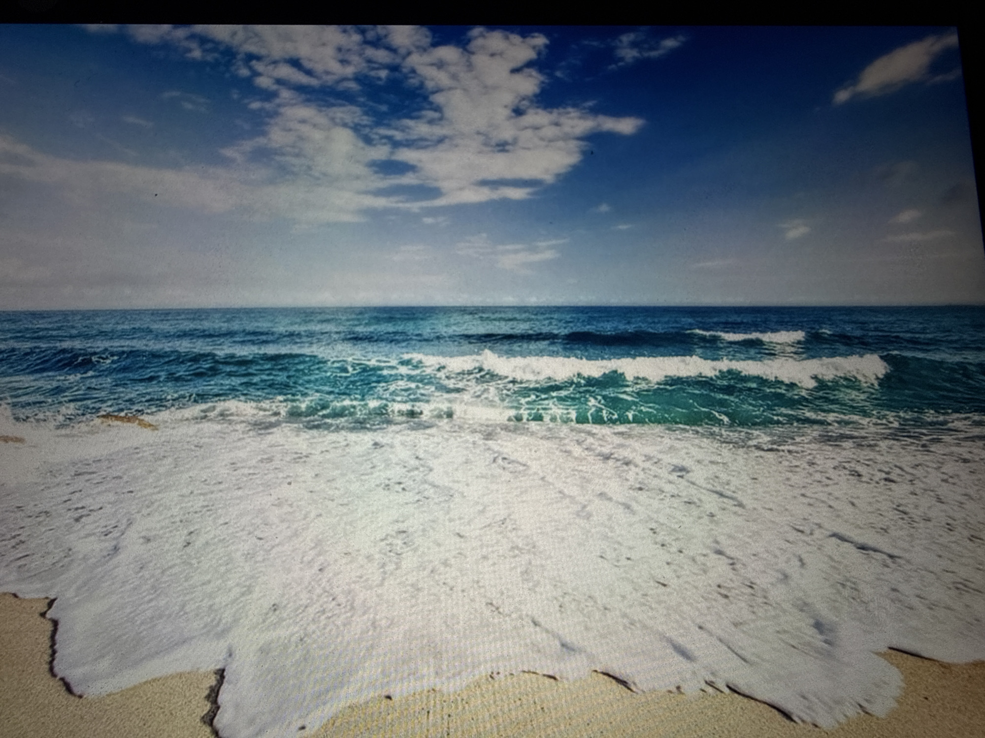 A photograph depicts a serene beach scene. The sky is a vibrant blue, dotted with fluffy white clouds, which gradually transition to a hazy lighter shade towards the horizon. The ocean is a deep turquoise, with white-capped waves rolling towards the shore. The foreground is dominated by a sandy beach, where foamy white waves are receding, leaving behind a wet sheen on the sand. The image captures the natural beauty of a coastline.