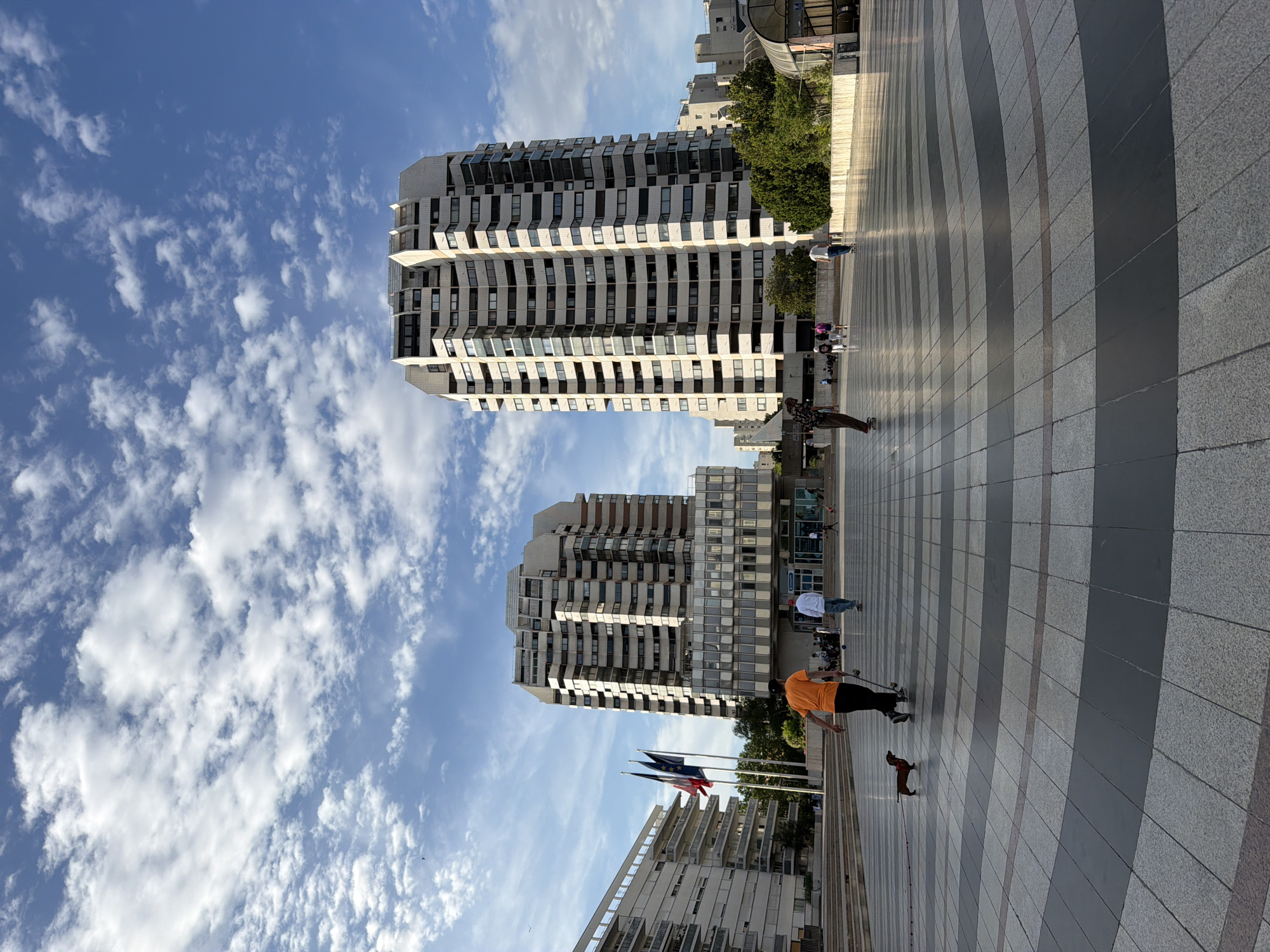 The image depicts a street scene in Créteil, France, under a bright blue sky with scattered white clouds, suggesting it's daytime and likely good weather. The perspective is from ground level, looking up towards modern, multi-story apartment buildings with many windows. Two prominent buildings dominate the view, featuring balconies and a repetitive facade design.
In the foreground, a paved walkway with a pattern of light and dark grey tiles stretches into the distance. Several people are present on the walkway, engaged in various activities. One person in an orange shirt is on a skateboard, while another, dressed in dark clothing, is walking a small dog. Further back, a person in a white shirt appears to be walking, and a group of people are gathered near the entrance of one of the buildings. Trees are visible between the buildings and along the edge of the walkway, adding a touch of greenery. Three flags are mounted on poles in the middle ground, indicating a public space. The overall atmosphere seems relaxed and urban.