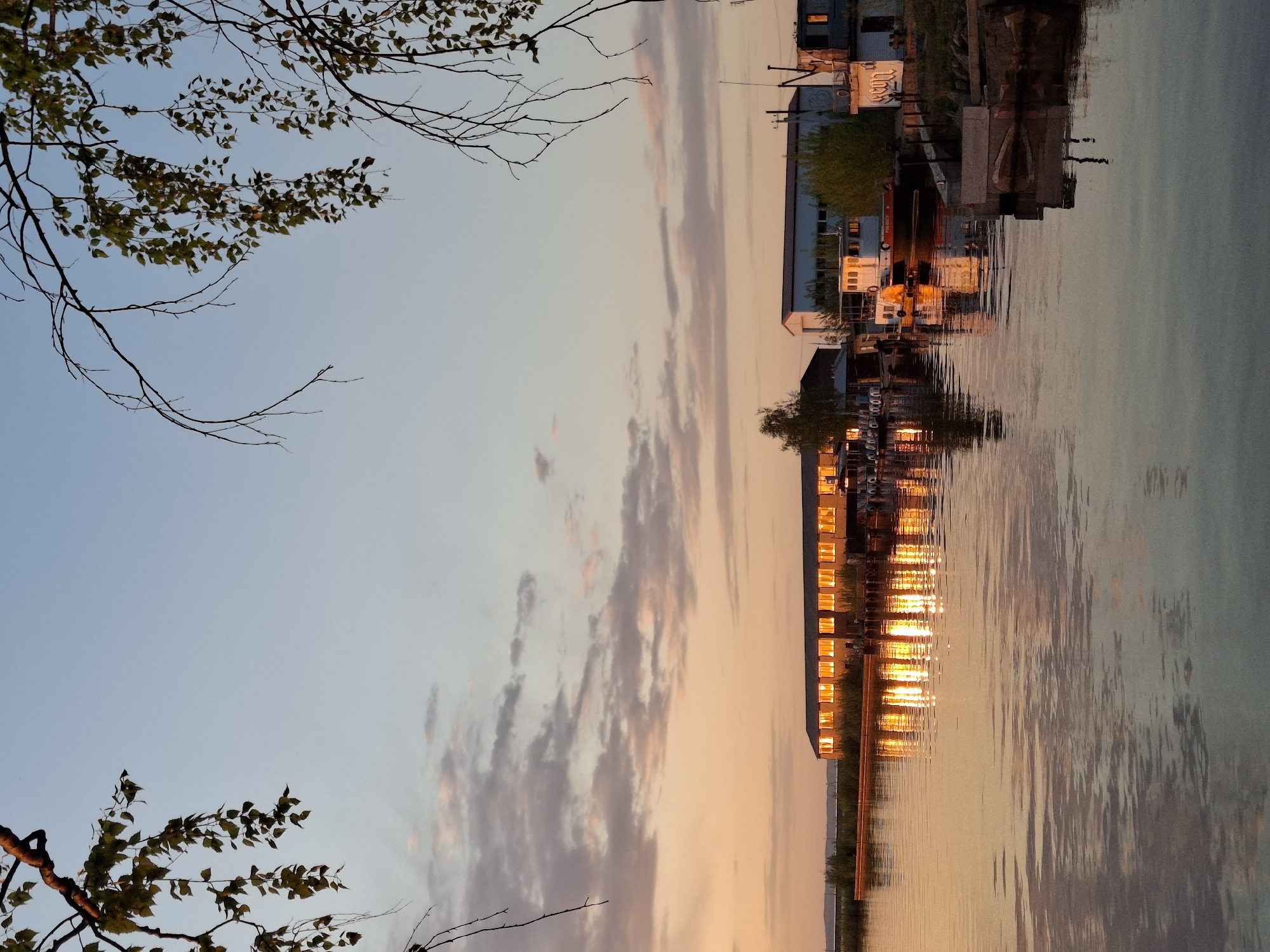 The image depicts a waterfront scene in what is likely the city of Chaikovsky, Russia, at twilight or dusk. The primary subject is a body of water, reflecting the buildings and sky above.
**Subjects & Objects:**
* A multi-story building or structure is visible, with numerous windows illuminated by golden light.
* Boats, machinery, and other industrial equipment can be identified in the area near the water.
* Tree branches and leaves are in the foreground, appearing silhouetted against the sky.
* The sky is a mix of light blue and orange tones, indicating either sunrise or sunset.
**Environment:**
* The environment appears to be a built-up waterfront area, possibly an industrial port or harbor.
* The body of water is relatively still, allowing for clear reflections of the surrounding buildings and sky.
**Actions & Interactions:**
* The lights within the building are turned on, suggesting activity inside.
* The water reflects the light and colors of the sky and buildings.
**Details specific to Chaikovsky:**
* Given the presence of what seems like a heavy industrial and waterfront area, it's likely that this area is the shores of the Votkinsk Reservoir on which Chaikovsky is situated. This is speculative, because there are few unique identifying objects to verify the location.