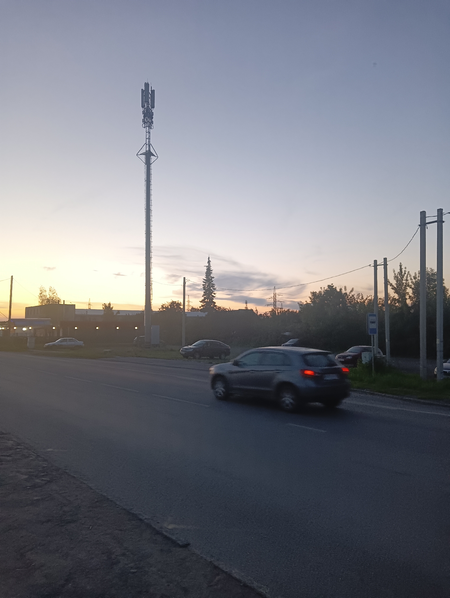 The image depicts a telecommunication tower and a road scene at dusk in Kamensk-Uralsky, Russia. A tall, lattice telecommunication tower stands prominently against a pale sky transitioning from yellow to blue. In the foreground, a paved road stretches into the distance. Several cars are visible on the road, some in motion, indicated by motion blur. One car, a dark grey SUV, is in the process of overtaking another vehicle. On the side of the road, there are trees, utility poles with wires, and a bus stop sign. A low-rise building with a light-colored facade is situated behind the road and to the left of the tower. The overall atmosphere suggests an urban or semi-urban environment during the evening hours.